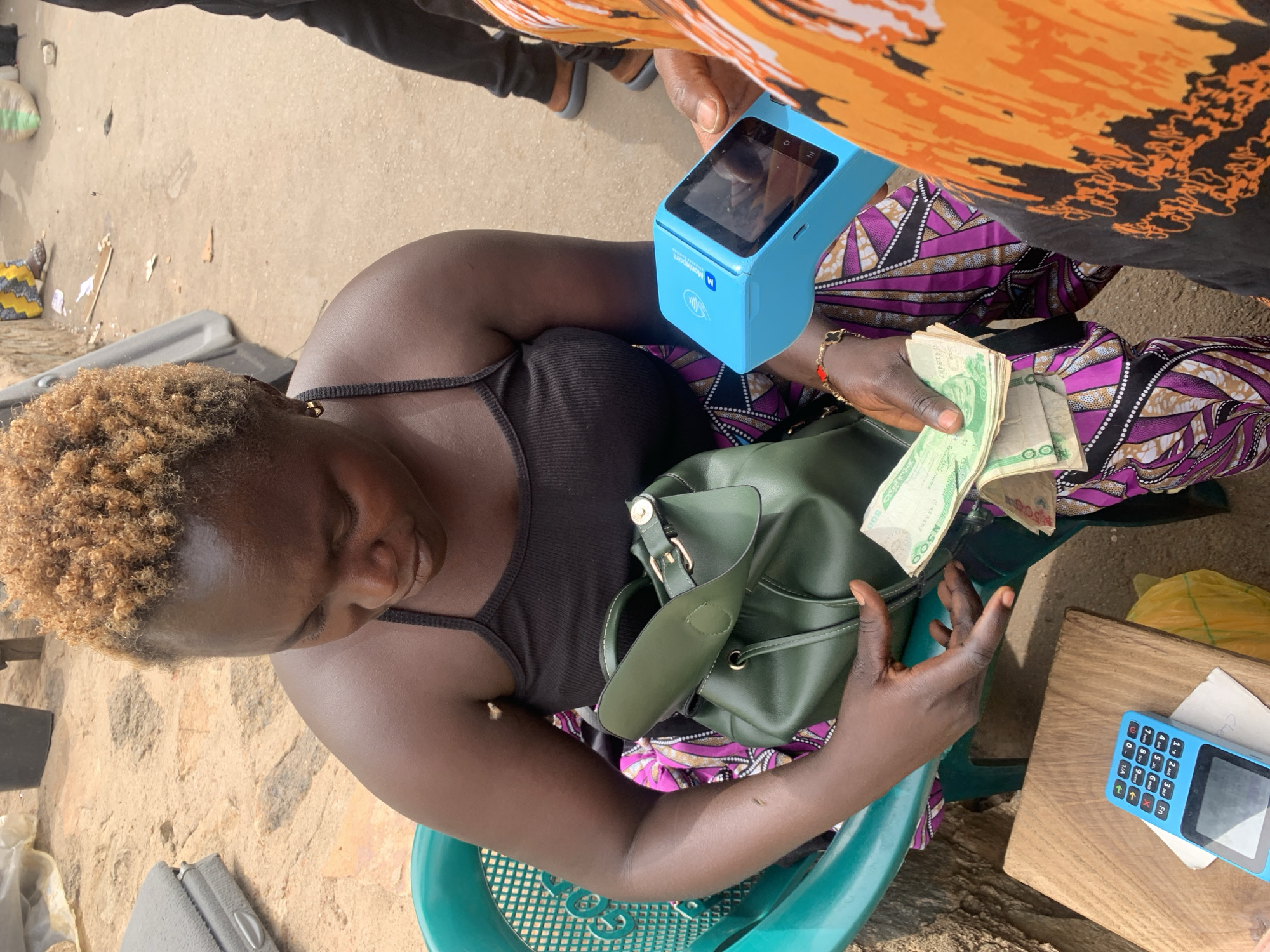 A woman with short, blond-tinged hair is seated outdoors, likely in Bukuru, Nigeria, given the currency and context. She is wearing a black tank top and sitting on a teal plastic chair. She holds a stack of Nigerian Naira notes (500 Naira denomination visible) in her right hand. Her left hand appears to be reaching into or adjusting a dark green leather-like handbag. A person with orange and black patterned clothing is standing beside her, holding a light blue Moniepoint mobile point-of-sale (POS) device. Another blue POS terminal is on a small wooden table beside the woman. The ground appears to be concrete or a similar rough surface. There is other miscellaneous debris and items scattered around the area.