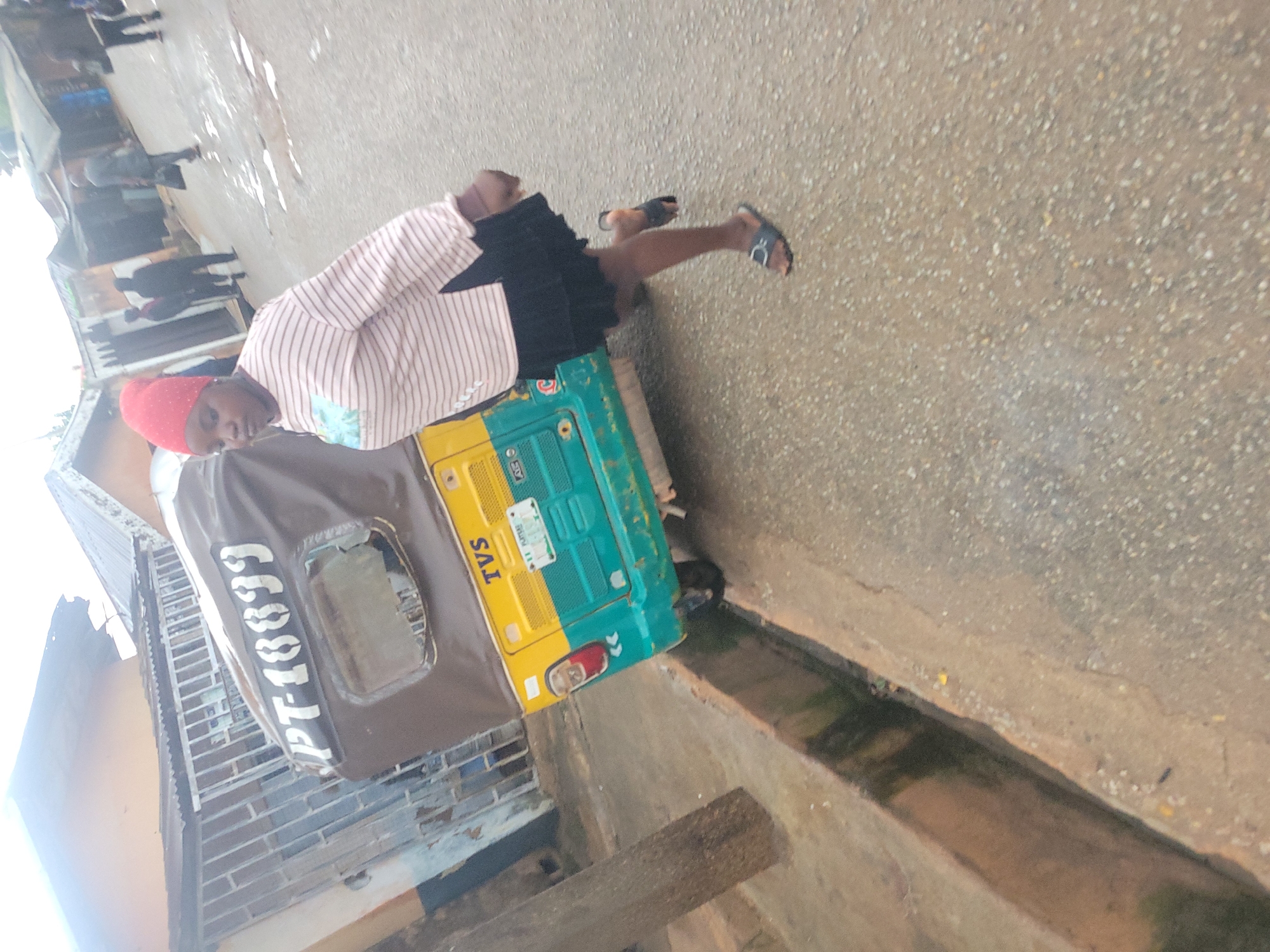 A woman wearing a red headscarf and a striped long-sleeved shirt walks past a three-wheeled vehicle, possibly a tuk-tuk, in Jos, Nigeria. The vehicle is labeled with "PT-18899" and "TVS." The ground appears wet, with puddles of water visible, suggesting it may have recently rained. Buildings and other people are visible in the background, indicating an urban setting. The woman is in motion, with one leg raised as she walks. The overall scene depicts a slice of everyday life in a Nigerian city.