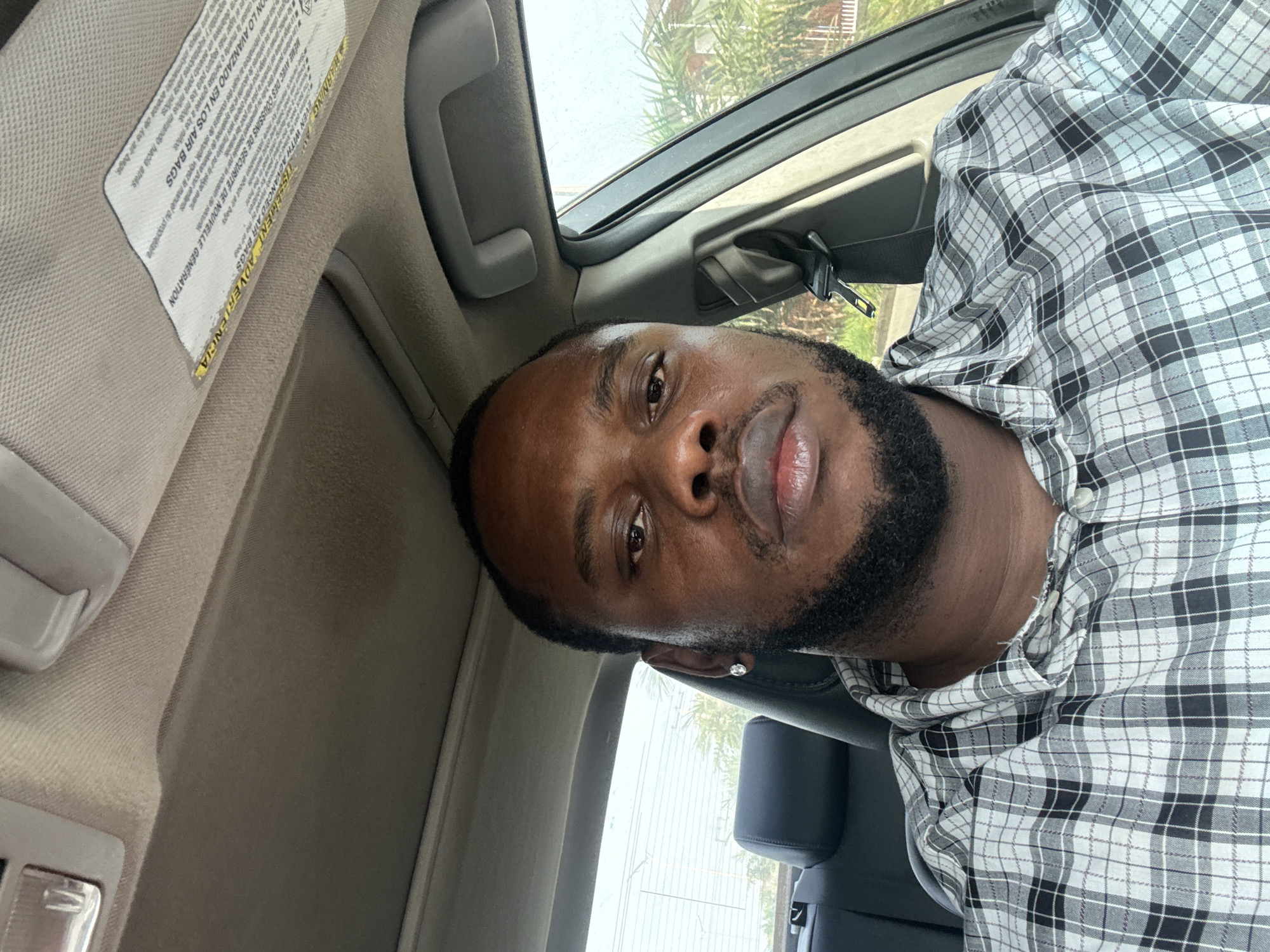 A medium-shot, indoor self-portrait of a Black man, apparently seated inside a vehicle. The man has a dark beard, short hair, and is wearing a patterned shirt. He appears to be in the driver's seat. The interior of the car is visible, including the roof lining, sun visor, and a glimpse of the window showing buildings and trees outside. Based on the available image data, it's impossible to pinpoint the precise location in Lugbe, Nigeria or confirm any specific actions or interactions taking place.