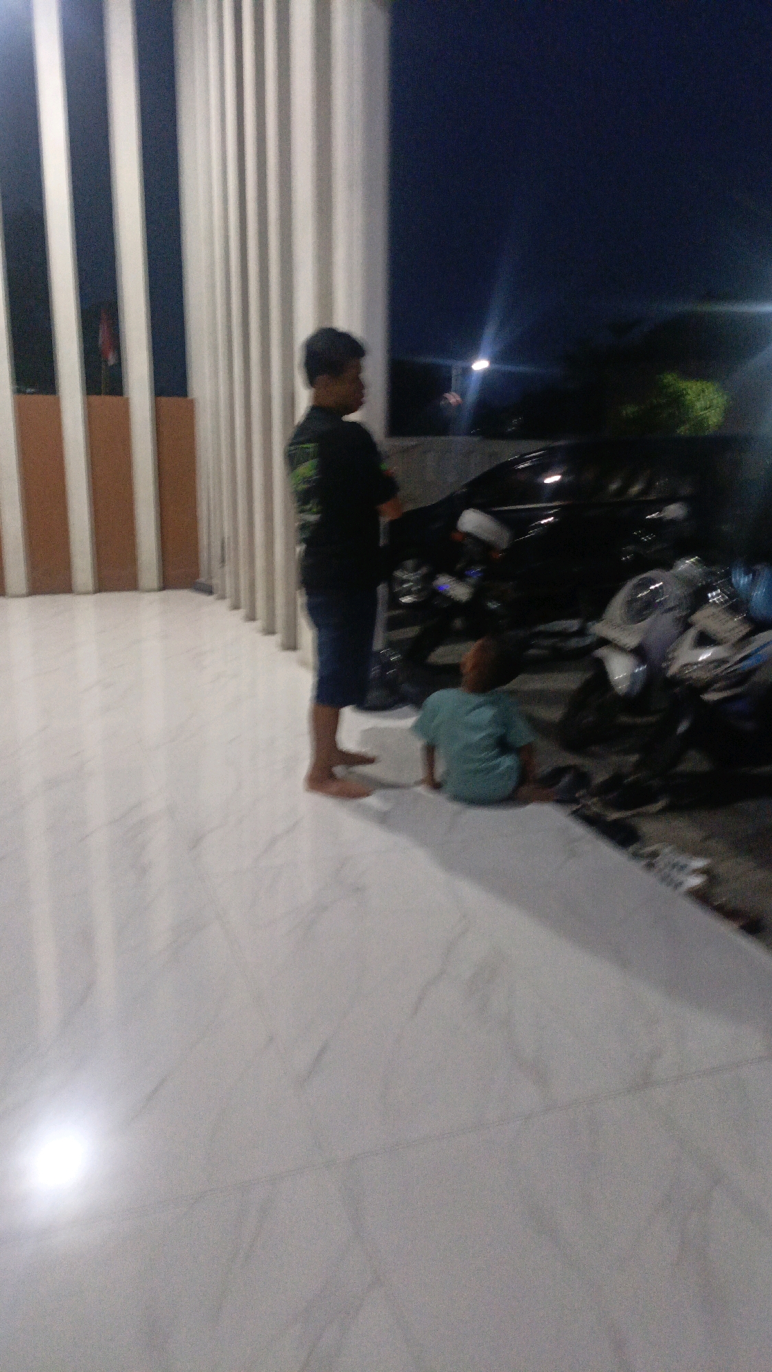 In the city of Ngaru Aru, Indonesia, a low-angle, nighttime shot depicts a scene with a man and a child standing near several motorcycles. The man, facing away from the camera, is dressed in a dark t-shirt and blue shorts, with bare feet. A child, facing away from the camera, is seated on the ground wearing a light blue t-shirt. Behind them, a row of white vertical pillars with darker brown sections stands against a dark blue night sky. Several motorcycles are parked in the background. The ground is paved with large, light-colored tiles. A bright light source illuminates the floor on the left side of the frame.