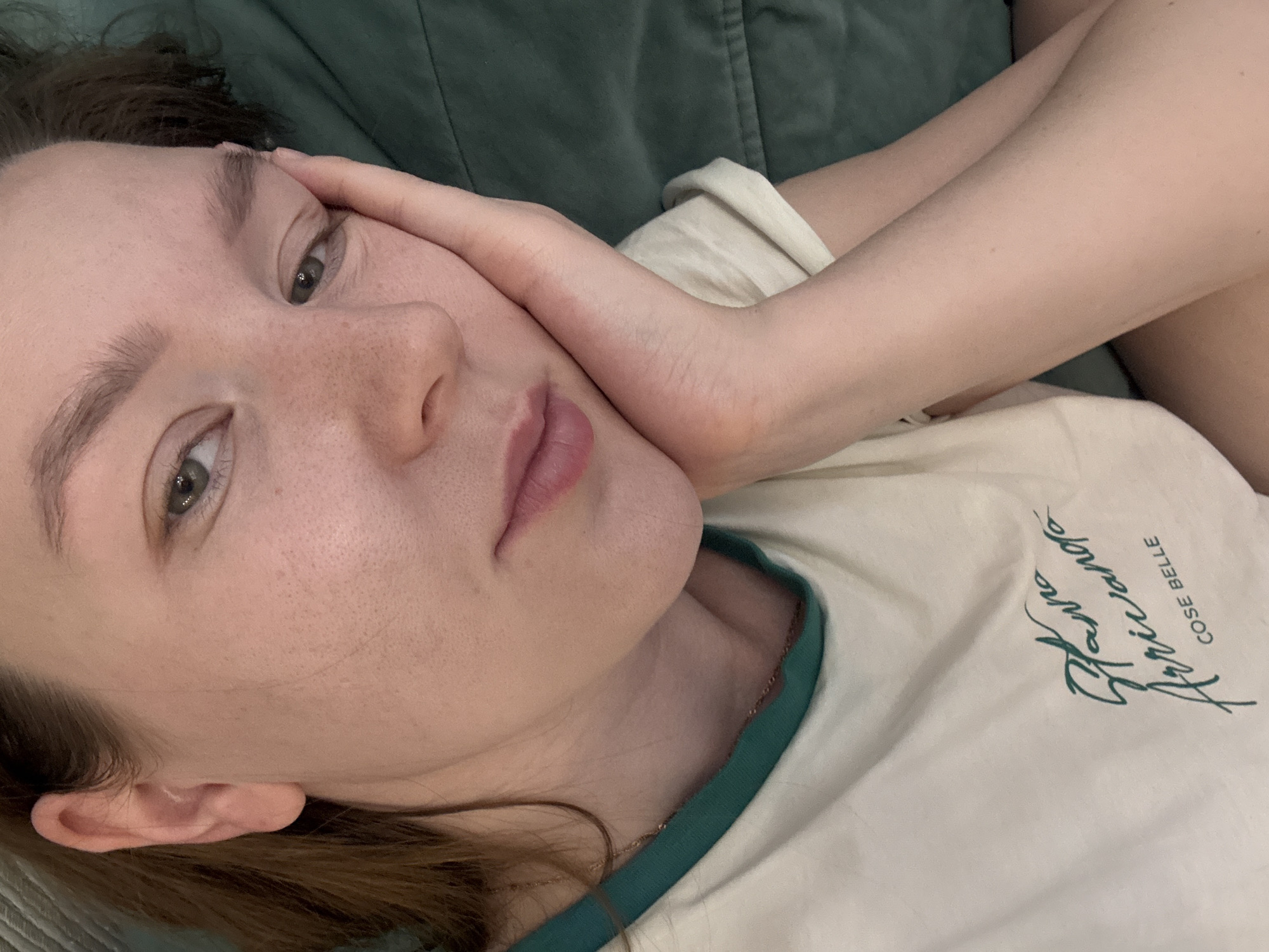 A woman is lying down, resting her head on her hand. She is wearing a white t-shirt with green trim and a cursive design. She has fair skin, light brown hair, and visible freckles. The background is a green, textured surface, possibly a couch or pillow. The scene is an indoor setting. There is no evidence suggesting the location is Izhevsk, Russia.
