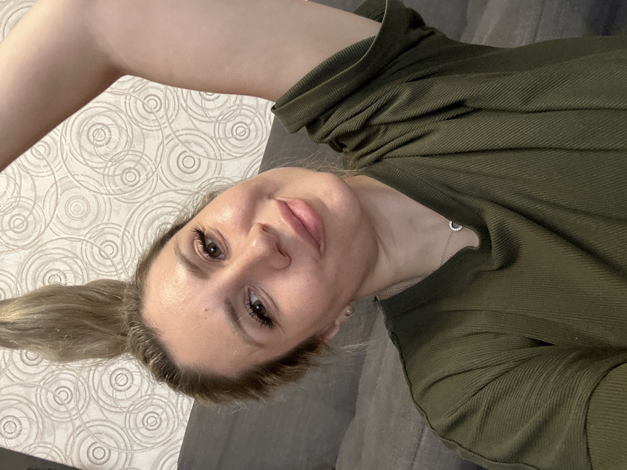 A woman with blonde hair pulled into a high ponytail is looking into the camera. She is wearing a green top and a silver necklace. Her left arm is raised above her head. The background is a wall with a circular pattern. The scene appears to be indoors, likely a living room, given the presence of a sofa. The exact location in Novoaltaysk, Russia cannot be determined from the image.