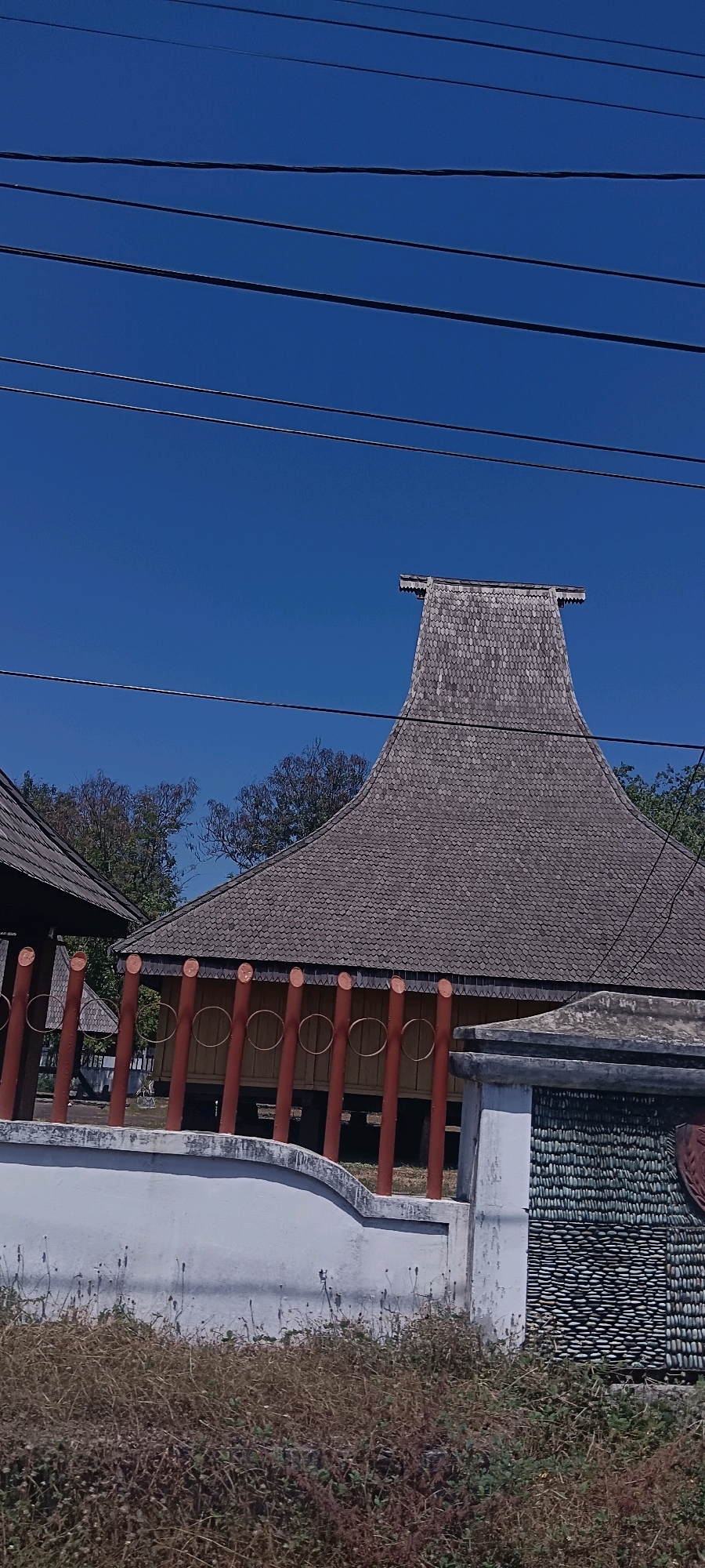 This is a medium shot, eye-level photograph captured on a bright, clear day, showcasing a traditional structure with a distinctive, upswept roof in the city of Ende, Indonesia. The roof, covered in overlapping wooden shingles, dominates the upper portion of the frame and tapers to a sharp point at its apex. The building appears to be a traditional house or hall, characterized by its stilts and ornate, red-painted wooden pillars supporting a covered walkway. A white wall, adorned with decorative stonework, runs along the foreground, partially obscuring the lower part of the structure and the surrounding dry, brown vegetation. Several power lines traverse the bright blue sky, creating a visual contrast with the natural and architectural elements below. The overall impression is one of cultural heritage set against a backdrop of everyday utility lines.