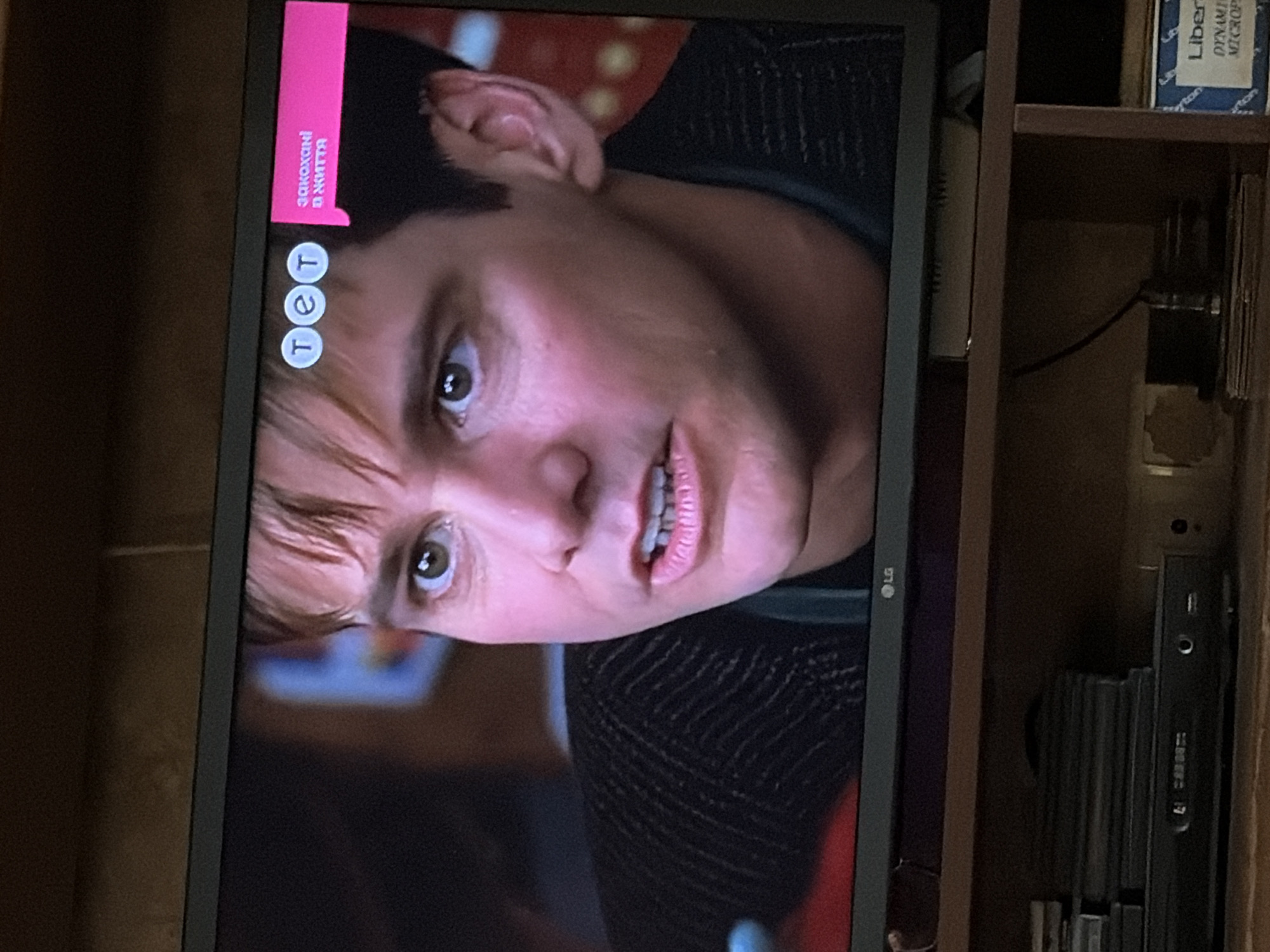 The image shows a close-up of Jim Carrey's face on a television screen. He is looking to the side with a concerned expression, his mouth slightly open, revealing his teeth. He has light brown hair, fair skin with visible pores and some redness, and green eyes. He is wearing a dark, textured sweater. In the upper right corner of the screen, there are two circular logos with the letters "TET" and a pink rectangular banner with Ukrainian text that translates to "Loved in life." The television is positioned on a dark wooden shelf unit, below which are various electronic devices and stacked items. The overall environment appears to be an indoor setting, likely a living room.