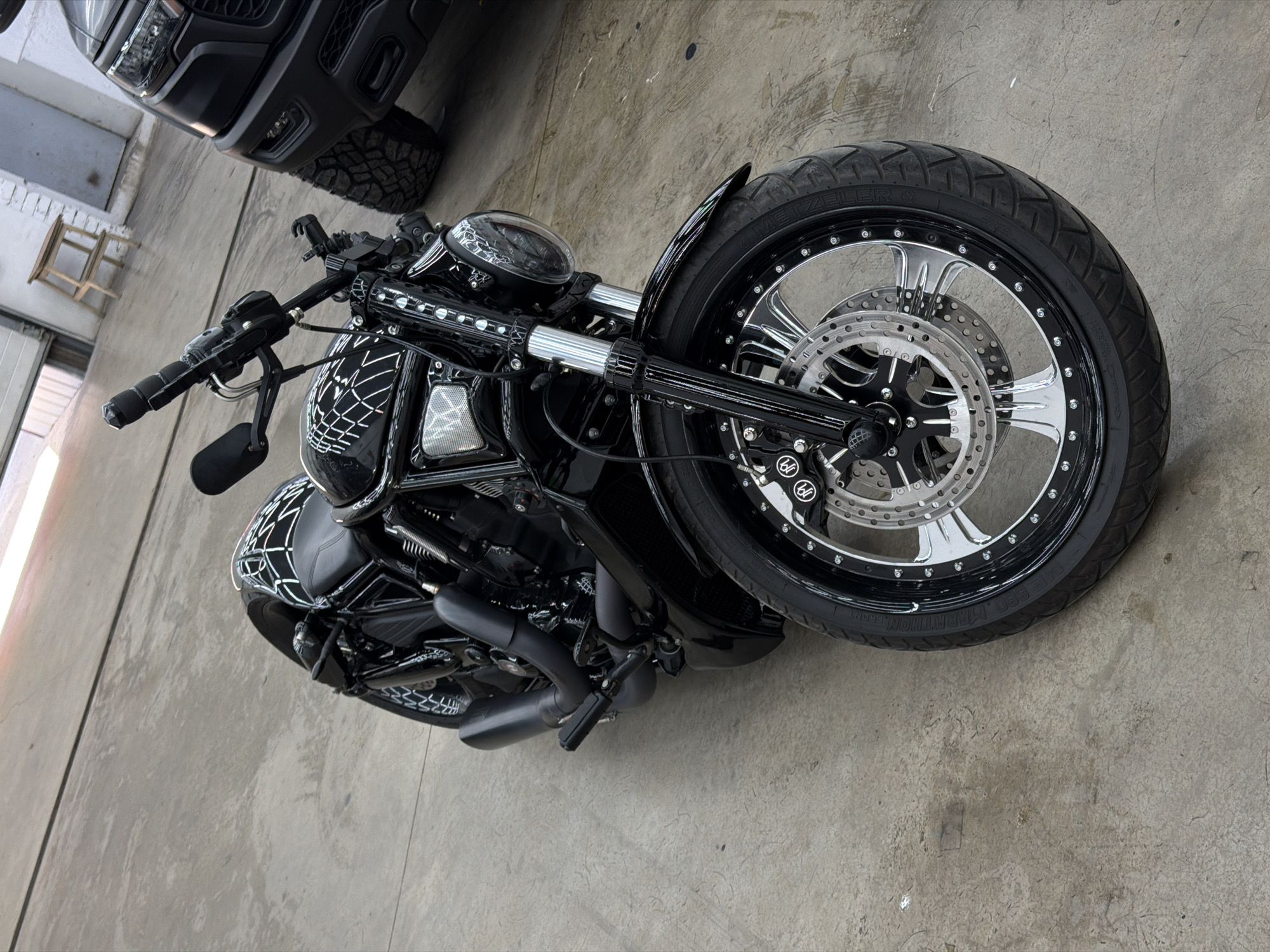 An overhead shot captures a highly customized black motorcycle parked indoors, likely in a garage or showroom in Krasnoyarsk, Russia. The motorcycle, viewed from above and slightly to the front, dominates the frame, showcasing its intricate details against a plain concrete floor.
The motorcycle features a glossy black finish with distinctive white or silver spiderweb-like graphics adorning the fuel tank and rear fender, adding a unique, edgy aesthetic. Its robust front wheel is a prominent feature, fitted with a thick tire and a striking black and chrome multi-spoke rim, secured with numerous small bolts around its edge. Dual disc brakes with drilled rotors are visible on the front wheel, and the brake calipers bear a stylized "6b" or "bb" logo. The tire sidewall clearly displays the brand "METZELER" and partially "MARATHON" and "FRONT". The front forks are chrome, contrasting with the black handlebars equipped with mirrors.
In the background, a portion of a black SUV or truck is visible, with its front bumper, grille, and a large, aggressive tire peeking into the frame. Further back and to the left, a simple wooden stool can be seen against a light wall, suggesting a utilitarian or workshop environment. The concrete floor exhibits some minor stains and markings but is generally clean.
The scene depicts no activity; the motorcycle is stationary. The lighting is even and diffused, indicative of an indoor setting, possibly with artificial lights or soft natural light from unseen windows. No specific time of day is discernible, and no emotions are conveyed. The motorcycle's exhaust pipes are short and black, matching the overall dark theme, and a small "W" logo is visible on the front wheel hub.