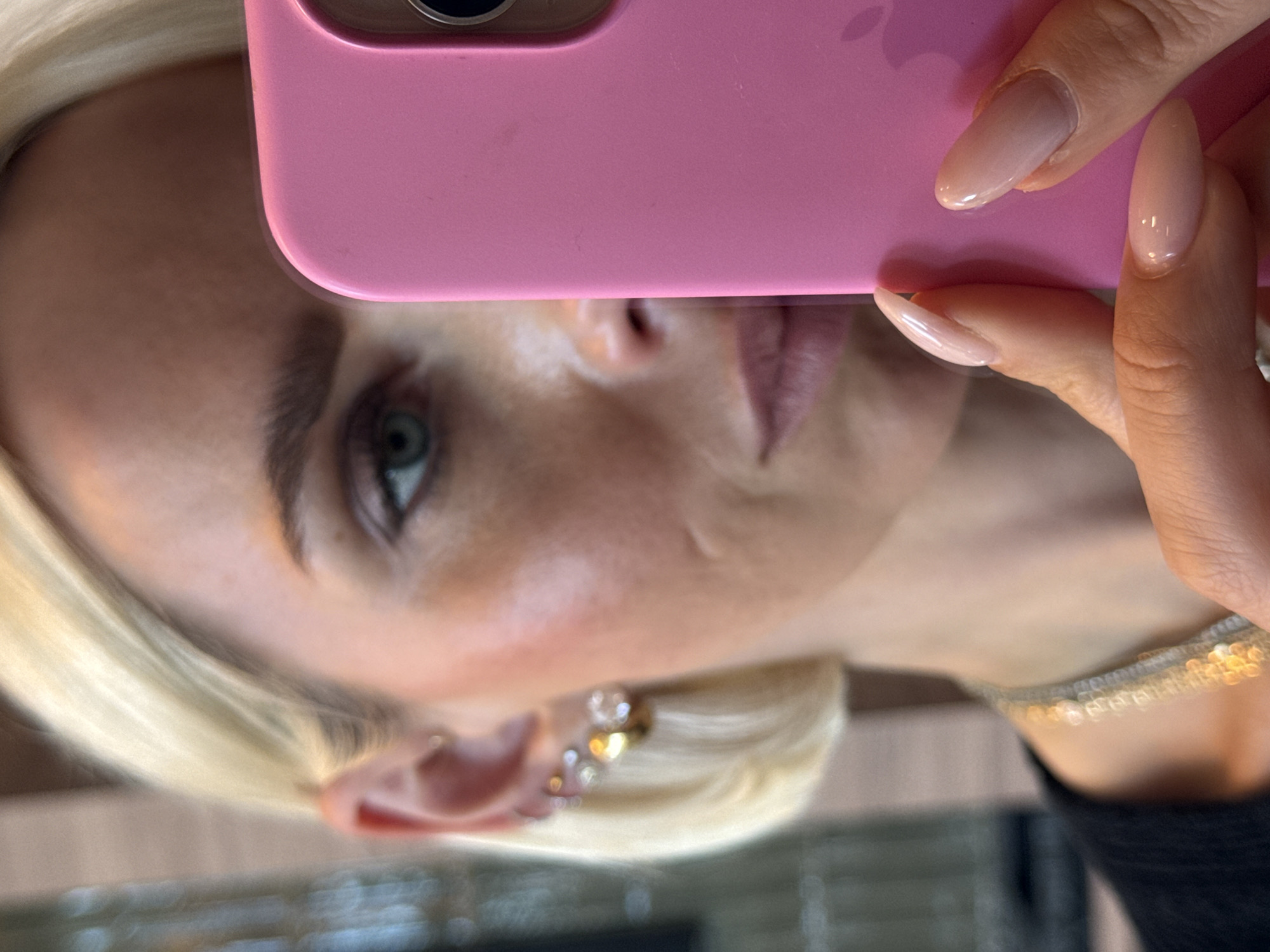 A close-up image depicts a person with blonde hair and fair skin holding a pink phone up to their face, obscuring the upper portion of their head. Only one green eye is visible. The person has an earring with multiple gold colored adornments. They are wearing a silver and gold necklace. The location in San Diego, United States of America is not discernible from the information presented in the image.