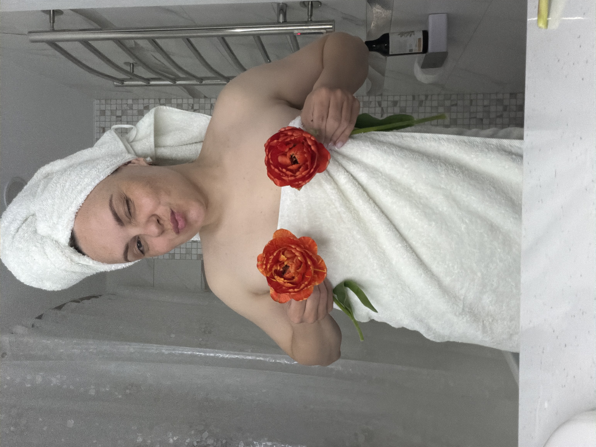 A person is depicted in a bathroom, likely after showering or bathing, in what is contextually identified as Votkinsk, Russia. The subject, appearing to be an adult female, is wrapped in a white terry cloth body towel, with a matching towel turbaned on her head. She holds two vibrant orange and red double tulips, one in each hand, positioned across her chest. Her expression is playful, featuring one eye partially closed in a wink and lips slightly pursed.
The background reveals a light-colored tiled wall, including a horizontal band of small square mosaic tiles. Above this, a chrome heated towel rack is mounted. To the upper right, a dark-colored bottle, possibly wine, with an indistinct label, rests on a ledge next to a small white rectangular wall fixture. To the far left, a translucent shower curtain or screen is visible, while a wet, possibly glass, shower enclosure with water droplets is seen on the right. The lighting is bright and even. The only visible text, found on the bottle label, is too blurry to be read clearly.