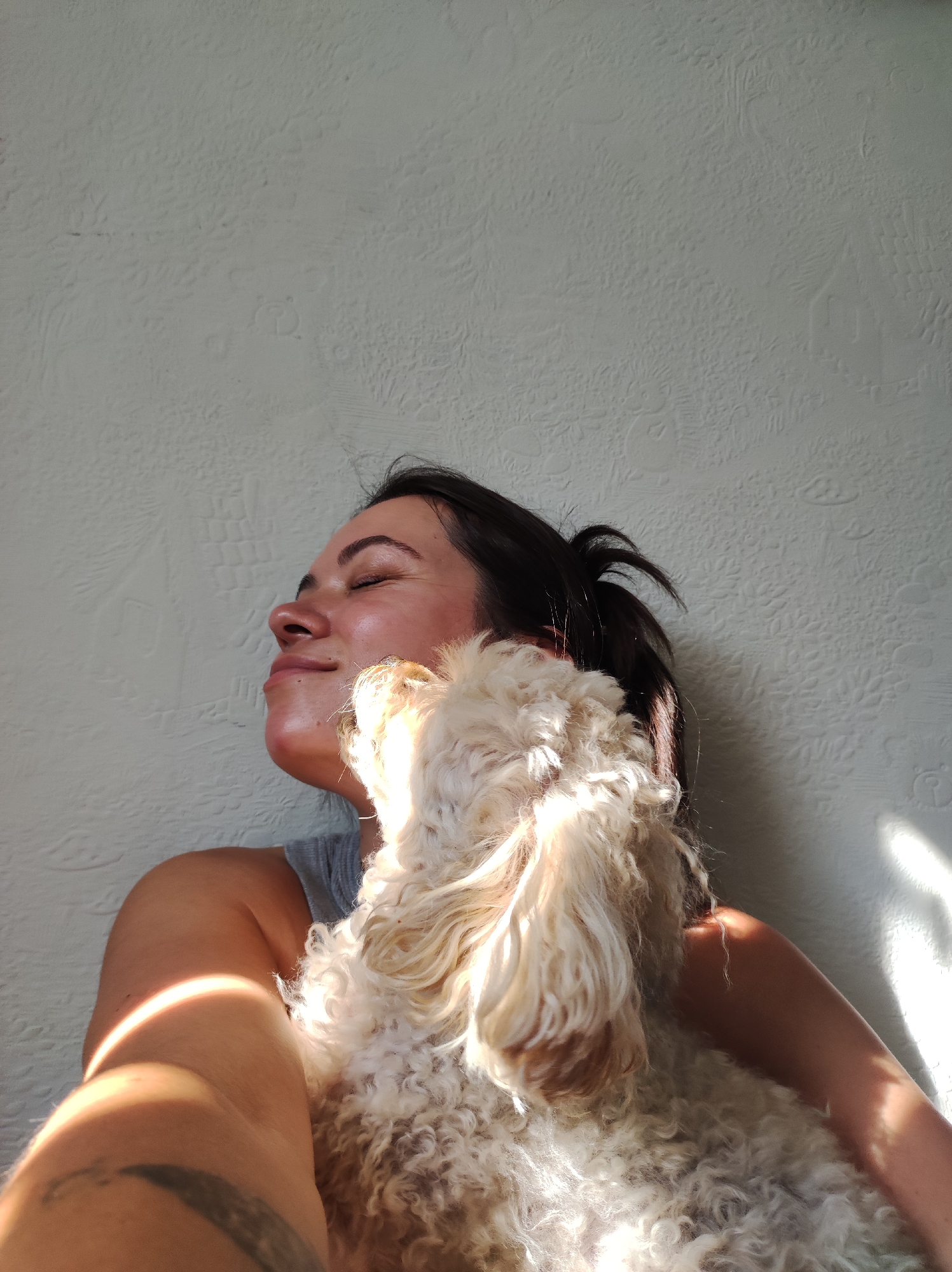 In the provided image, a woman is depicted with her eyes closed, her head tilted back and a contented expression on her face. She is embracing a fluffy, light-colored dog, which is nuzzling her face. The woman is wearing a grey tank top, and sunlight is casting a warm glow on her skin and the dog. The background is a textured, light-colored wall, with distinct patterns and shadows from sunlight. There is no visible indication of the city of Chaikovsky, Russia, within the image itself.