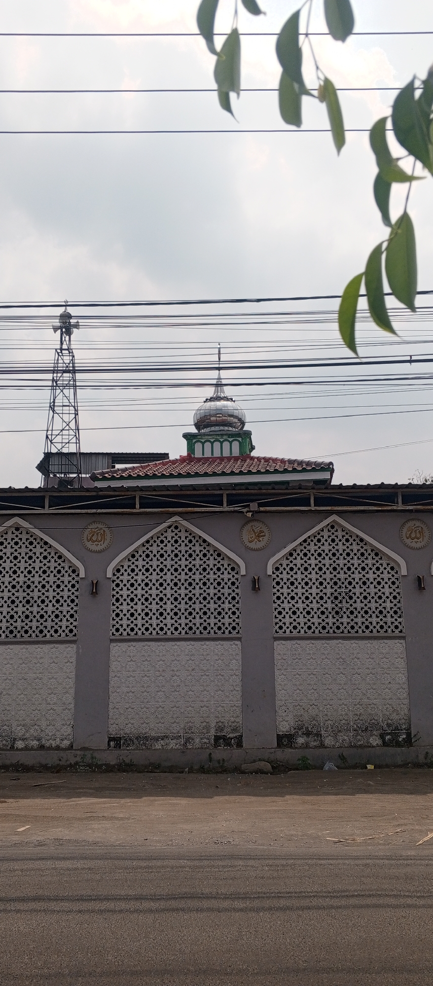 The image captures a frontal view of a mosque in Gulun, Indonesia. The mosque features a distinctive silver dome with green trim, situated above a red-tiled roof. A tall, lattice-like tower with speakers is visible to the left of the mosque. The mosque is partially obscured by a series of horizontal power lines that crisscross the frame. In the foreground, there is a gray concrete wall with decorative geometric patterns and Arabic calligraphy. A paved road and a patch of dirt are visible in front of the wall. The sky is overcast, with some clouds visible, and a few green leaves hang into the frame from the top right.