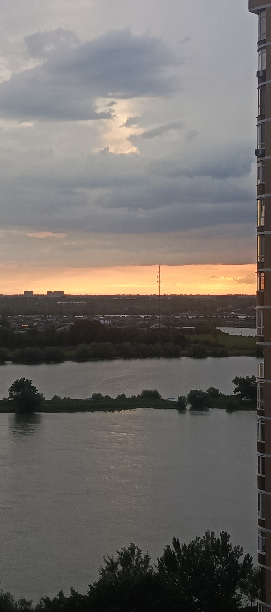 The image shows a river scene at dusk, with a modern apartment building on the right and a distant cityscape with buildings and a radio tower. The sky is cloudy with a warm orange glow from the setting sun peeking through. The river occupies the lower half of the frame, reflecting the muted light. A narrow strip of land with trees runs across the middle ground, dividing the river into two sections. The overall impression is one of a calm, urban landscape transitioning into evening. This scene is likely located in Krasnodar, Russia, given the typical architecture and riverine environment often found in the city.