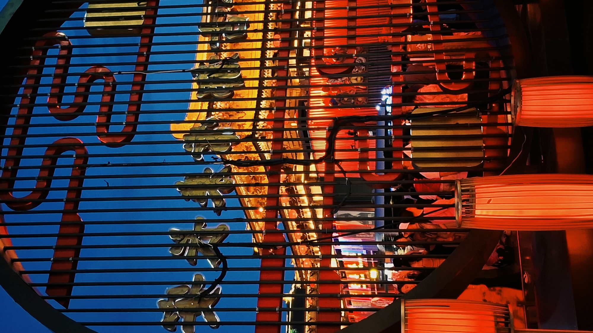 The image shows a section of a neon sign in Yanta District, China. The sign is predominantly red and gold with some blue visible in the background. It is viewed through a grating of horizontal bars. The red portions of the sign are tubular and stylized, while the gold elements are shaped like stylized flowers. To the right of the sign are red, cylindrical lanterns.