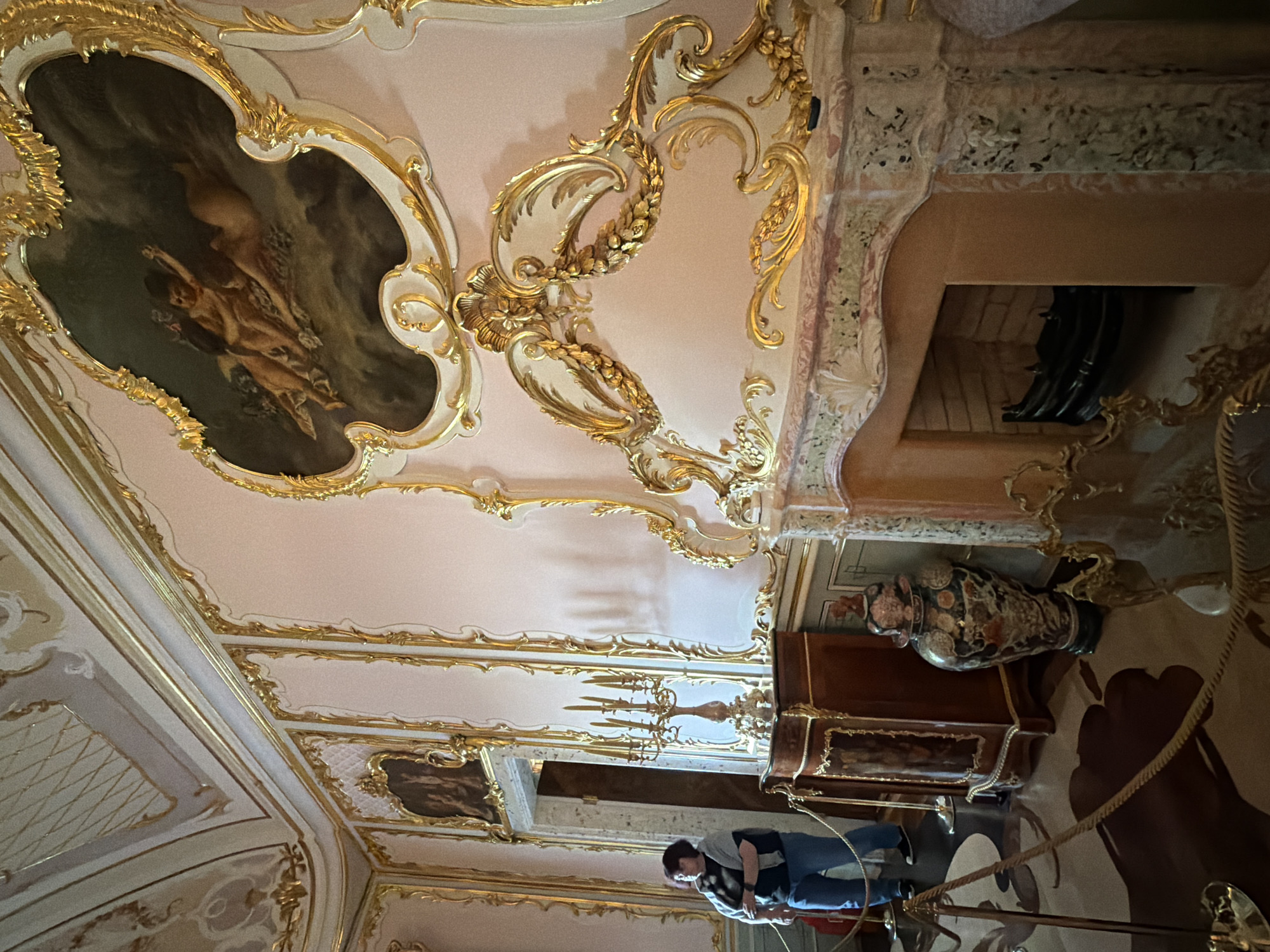 The image depicts a richly decorated interior of a room, likely a palace or museum, with ornate gold detailing on the walls and ceiling. A woman stands to the left, looking away from the camera, wearing jeans and a casual jacket. In the center of the room, a wooden cabinet with gold accents holds a candelabra. A large, decorative vase sits on the floor next to the cabinet. To the right, a marble fireplace with intricate gold trim is visible, with a black grate inside. A painting of cherubic figures is mounted on the wall above the fireplace, framed in elaborate gold. The floor appears to be covered in a patterned rug. Ropes and stanchions suggest that this is a public space, likely a historical exhibit.