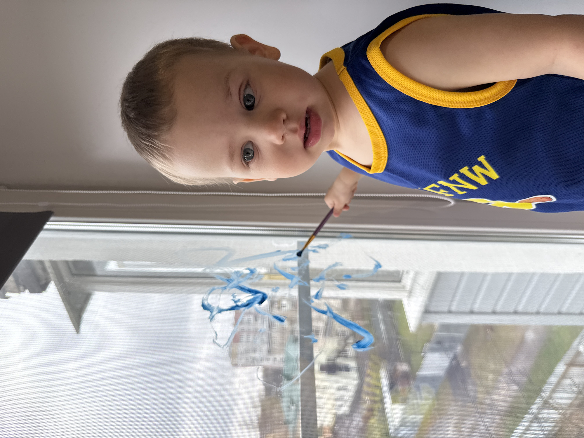 A young boy in a blue and yellow tank top is painting on a window. He is holding a paintbrush in his right hand and looking down at the window. The paint on the window is blue. The background shows a city view through the window screen. Location: Unknown, Unknown.