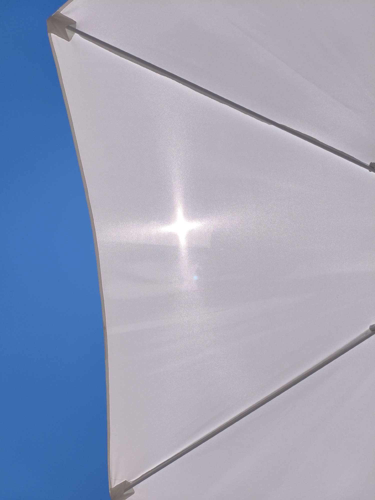 The image shows a close-up, low-angle shot of a white umbrella against a bright blue sky. The sun is directly overhead, shining through the fabric of the umbrella, creating a starburst effect and a lens flare. The umbrella appears to be a beach umbrella, made of a translucent white material stretched over a frame with visible metal poles. The bottom left portion of the image shows the curved edge of the umbrella, with the blue sky filling the rest of the frame. There are no people or any indication of a specific location within Sochi, Russia.