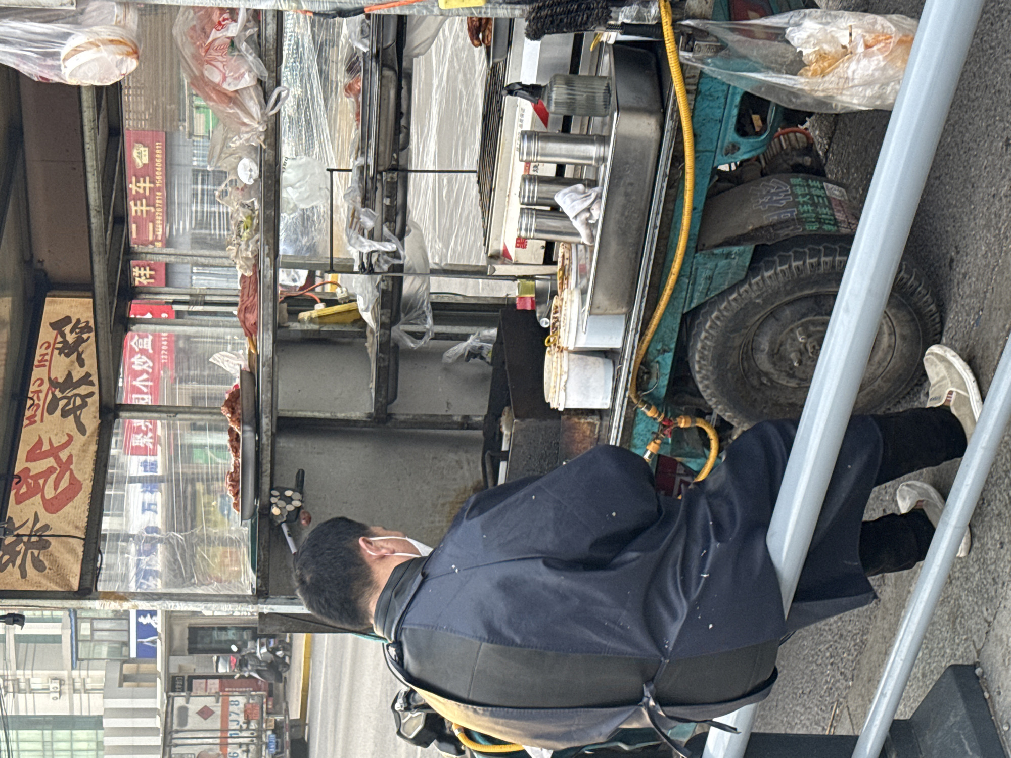 The image shows a street scene in Zhongtun, China, from an elevated perspective. A food vendor in a navy blue jacket is leaning against a gray metal structure with his back to the camera. He is wearing black pants and gray sneakers. His food cart is equipped with a cooking surface containing several metal cylinders. Various containers of food and supplies, many in clear plastic bags, are attached to the cart. Hanging signs with Chinese characters and advertisements for "Second Hand Cars" and "Electric Vehicles" are displayed on the cart. A street visible behind the cart includes buildings, parked scooters, and distant traffic.