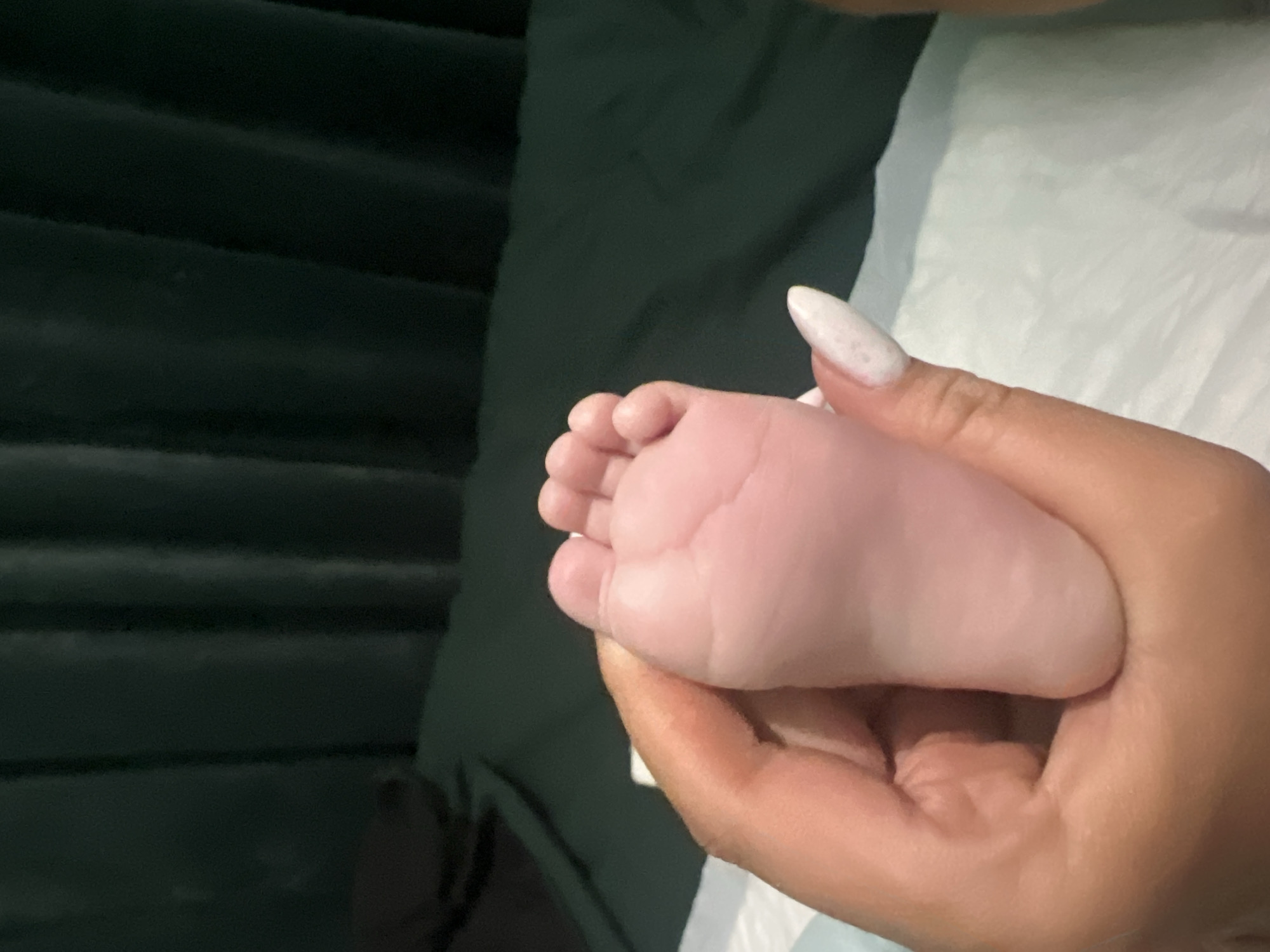 A human hand with long, white fingernails is holding a baby's foot. The background includes a dark green, possibly textured, surface and a white surface, possibly a cloth or sheet. The location cannot be determined from the image.