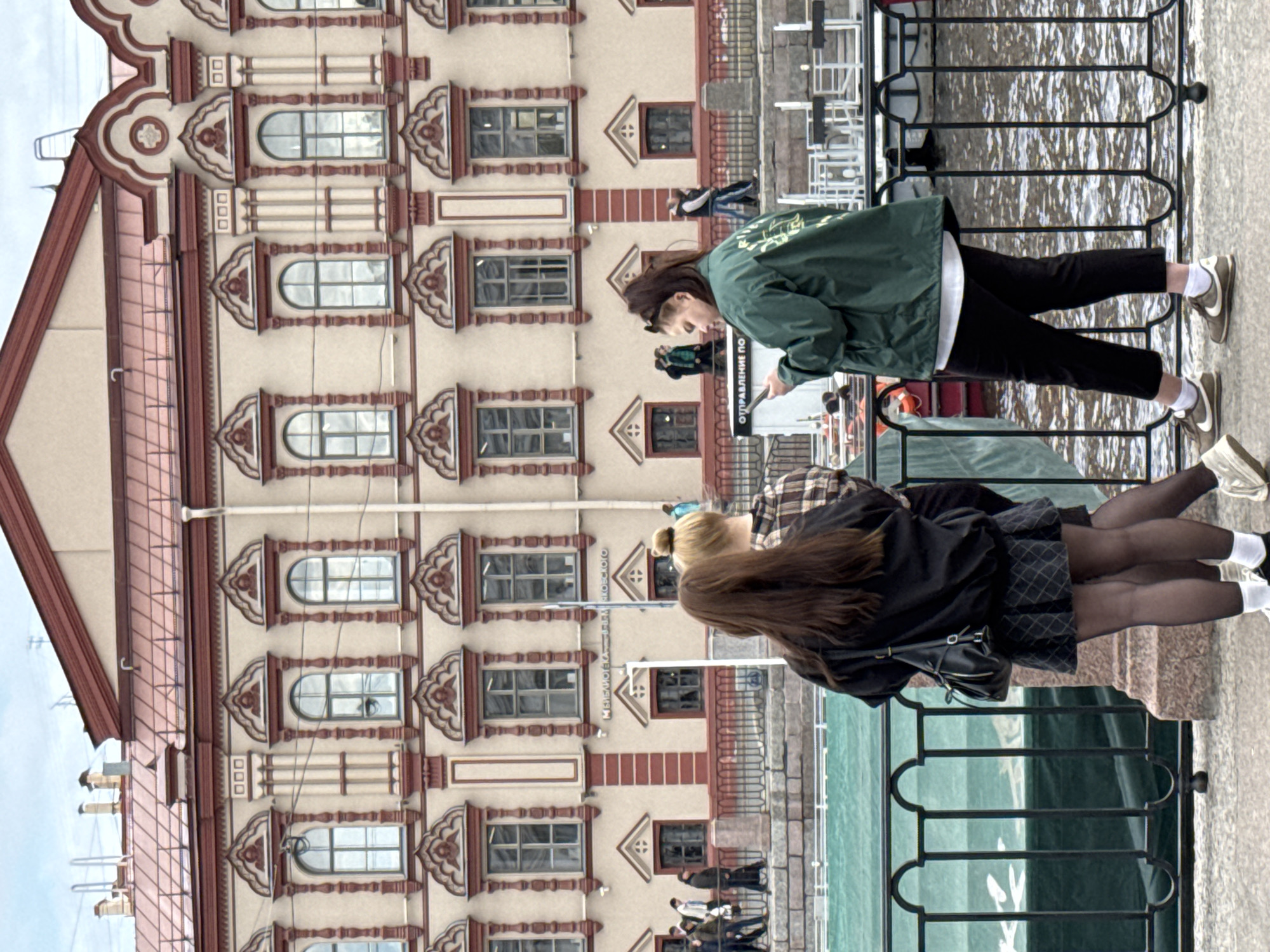 The image shows a group of people on a waterfront promenade in Saint Petersburg, Russia. In the foreground are two women standing near a black iron railing that separates the promenade from the water. One woman is wearing a green jacket and black pants. She is looking down and appears to be reading something. The other woman is wearing a black jacket, a dark patterned skirt, and black tights. Behind them is a building with decorative architectural details and multiple windows. In the background, there are other people walking along the promenade.