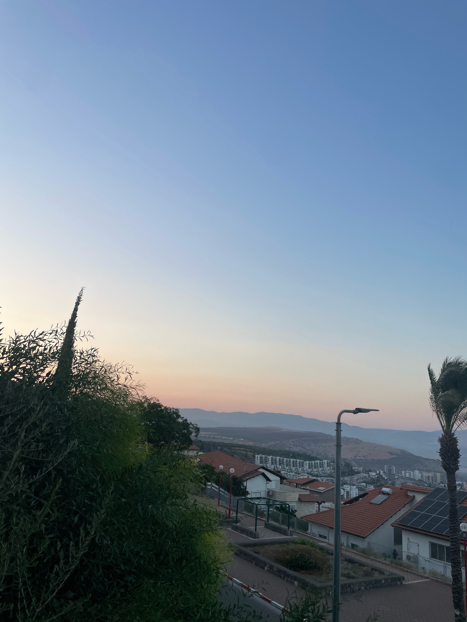 The image is an exterior, eye-level, landscape shot taken at sunset in Tiberias, Israel. The sky is a clear gradient of light blue to soft pink and peach hues near the horizon. In the foreground, a lush green tree with thin, elongated leaves dominates the left side of the frame. Behind the tree, a portion of a residential area is visible, characterized by buildings with orange tiled roofs. A tall, slender palm tree stands on the right side of the image, its fronds rustling. A street lamp with a modern design is positioned in the mid-ground, casting a shadow. The background reveals a hilly landscape with more buildings, some appearing to be apartment complexes and others single-family homes with solar panels on their roofs. The overall scene depicts a tranquil evening in a hillside town.