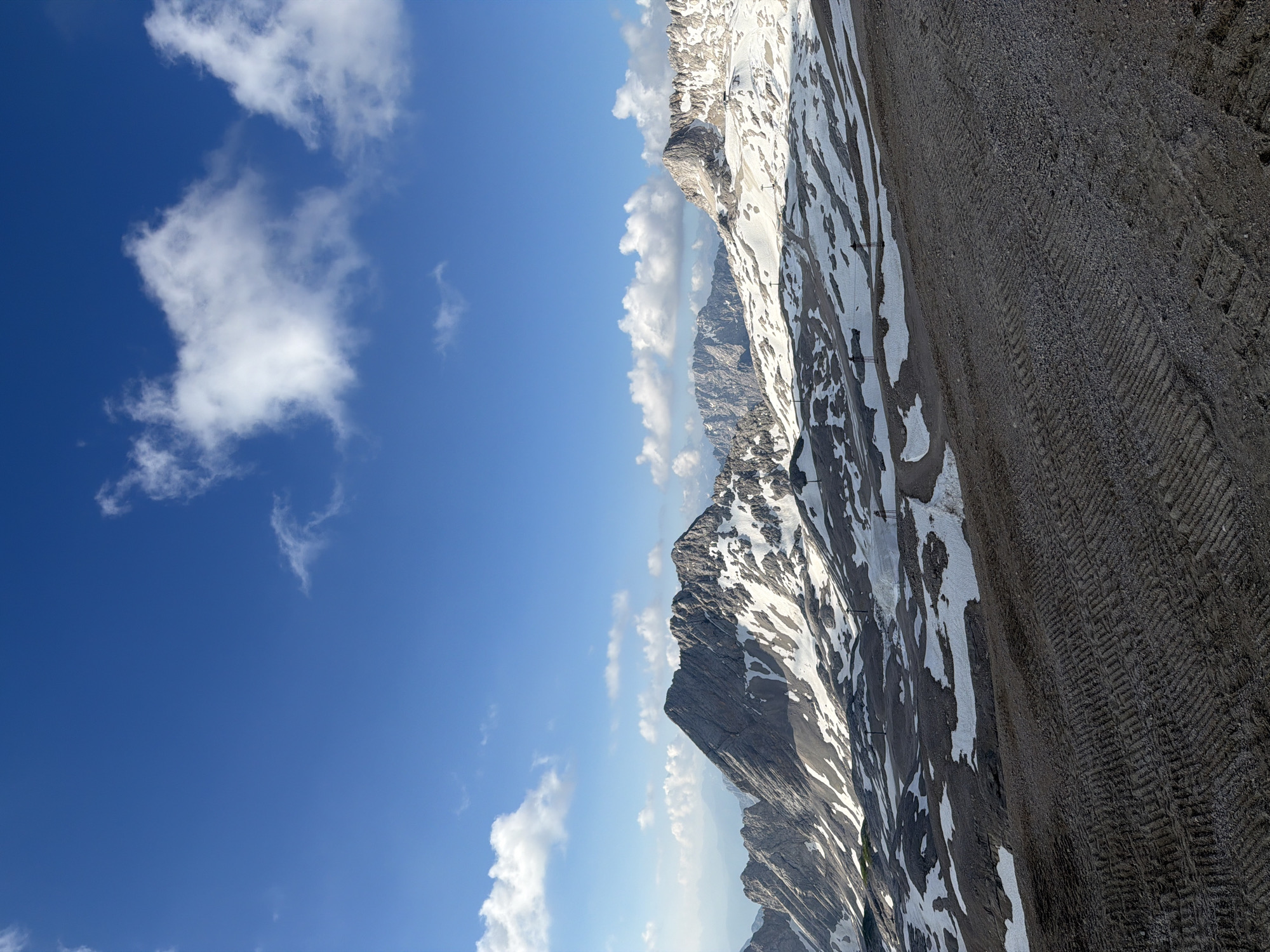 The image depicts a mountainous landscape under a bright blue sky with scattered white clouds. The foreground features a textured ground surface, possibly gravel or dirt, with distinct parallel track marks suggesting recent activity, such as from a vehicle or machinery. Beyond this, a vast, rugged mountain range stretches across the midground and background. Patches of snow are visible on the slopes, interspersed with dark rock formations. In the distance, further mountain peaks are visible, fading into a hazy blue. Several ski lift towers are sparsely distributed across the snowy slopes, indicating this might be a ski resort area. The scene is devoid of any people. This appears to be a high-altitude environment, characteristic of the Bavarian Alps, likely in or near Garmisch-Partenkirchen, Germany, given the presence of snow-capped mountains and ski infrastructure.