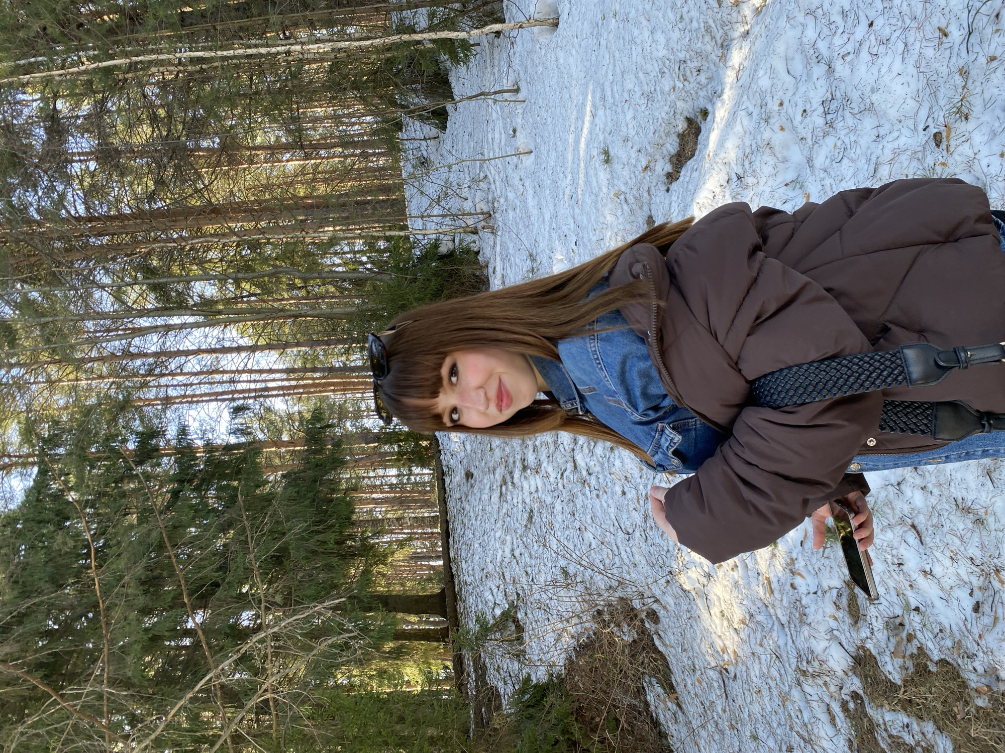 A young woman stands in a snowy woodland scene near Chaikovsky, Russia. She is wearing sunglasses on her head, a blue denim jacket under a brown puffer coat, and jeans. She holds a smartphone in her right hand. The ground is covered in snow with patches of dried grass and twigs visible. Behind her, a dense forest of tall, thin trees extends into the distance. The lighting suggests it is daytime, with the sun casting shadows through the trees.