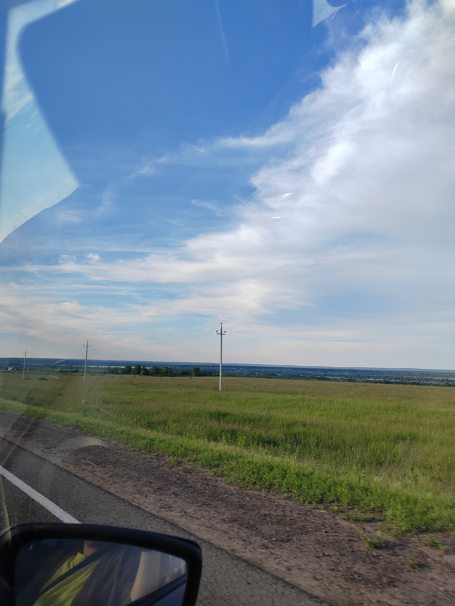 The image captures a rural landscape seen from the interior of a moving vehicle. The foreground features a paved road with a white solid line on its left edge, bordered by a dirt shoulder and an expansive green field of tall grass. In the bottom left, a portion of the vehicle's side mirror is visible, reflecting a person wearing a bright green or yellow garment.
The mid-ground is dominated by the vast green field, stretching towards a distant horizon. Several wooden utility poles, carrying power lines, punctuate the landscape, receding into the distance. A prominent pole stands slightly right of the frame's center. Beyond the field and poles, the horizon reveals a band of darker green trees, possibly indicating a distant forest or settlement, under a lighter, hazy blue sky.
The upper half of the image is filled with the sky, which transitions from a clear, bright blue on the left to an increasing presence of white, scattered clouds on the right. Wispy cirrus-like clouds are visible across the middle, and a larger, denser white cloud formation occupies the upper right quadrant. Faint, linear contrails are discernible in the sky. A significant reflection of the vehicle's interior, possibly a dashboard or window frame, is visible as a lighter blue, somewhat abstract shape in the top left corner.
The scene depicts a daytime setting, likely late morning or afternoon, under partly cloudy but bright weather conditions. The overall impression is one of open spaces and travel through a countryside environment. No explicit text is visible.