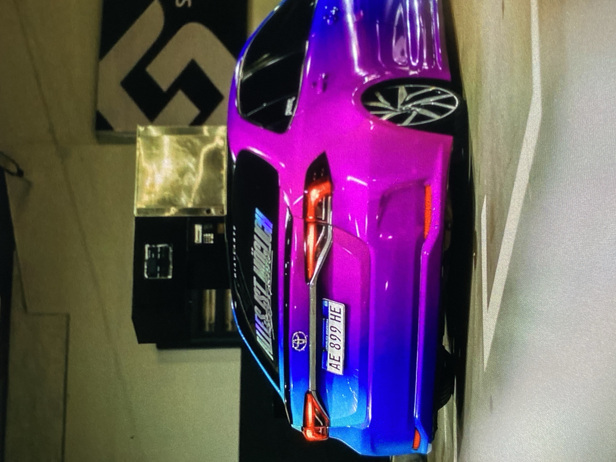 A modified, bright blue and pink Toyota car is parked in what appears to be a parking garage. The car's license plate reads "AE 899 HE." A sticker above the license plate is partially visible, reading "WEST ST. MICHEL". The car's modified rims are visible. The background includes a wall with a geometric black and white design and a vending machine. The location cannot be confirmed in the city of Lhokseumawe, Indonesia, based on the image alone.