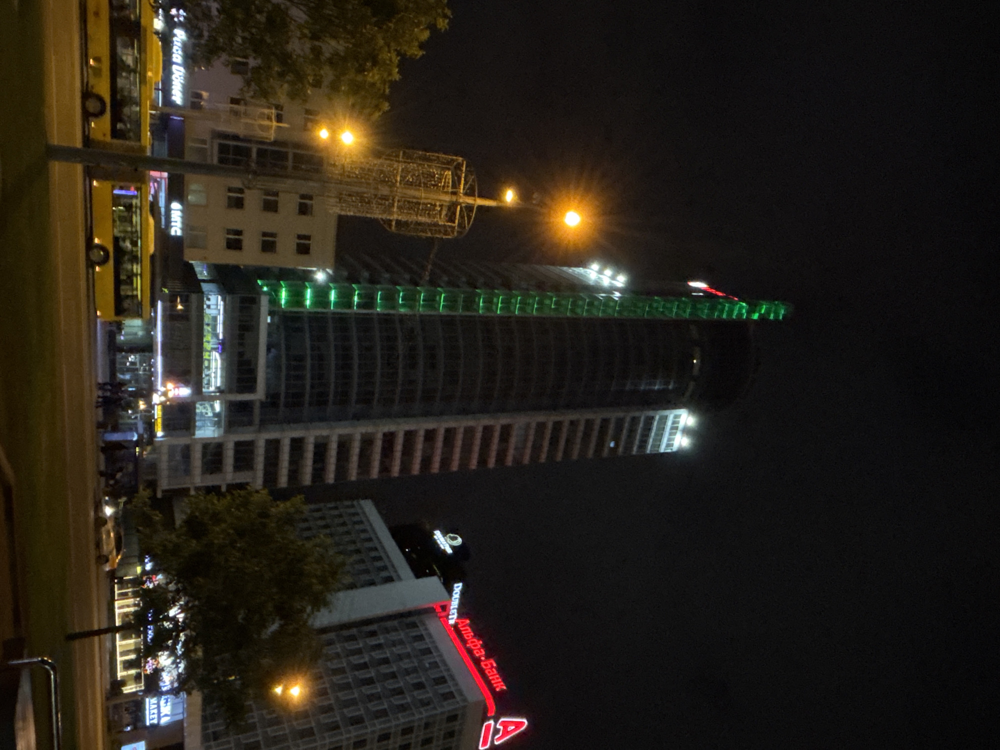This nighttime photograph depicts a street scene at Pobediteley Avenue 9 in Minsk, Belarus, featuring prominent commercial and hospitality buildings. The central focus is a tall, modern skyscraper, identified as the SkyLine Business Center, distinguished by vertical green LED light strips illuminating its left facade and bright white lights near its apex. To the right of the skyscraper stands the DoubleTree by Hilton hotel building, which displays a large, red neon sign reading "Альфа-Банк" (Alfa-Bank) and a smaller "DOUBLETREE" sign. A partially visible "DOUNUTS" sign is also present on this structure.
In the foreground, a wide urban thoroughfare, Pobediteley Avenue, is illuminated by numerous streetlights emitting a warm yellow glow, some of which exhibit prominent starburst lens flares. Two yellow public buses are captured in motion, driving from left to right across the frame. A light-colored sedan is also visible further down the road. On the left, a multi-story building features commercial signs at ground level, including "Pusa Doner" and "MTC." A tall street pole adorned with intricate, illuminated decorative elements stands in the mid-ground. Several indistinct human figures are visible on the sidewalks, particularly near the bus stop area. Trees with visible foliage are situated on the left and in the mid-ground, partially lit by ambient light. The sky is dark, indicating a clear night, and the scene is characterized by the artificial illumination of the urban environment.