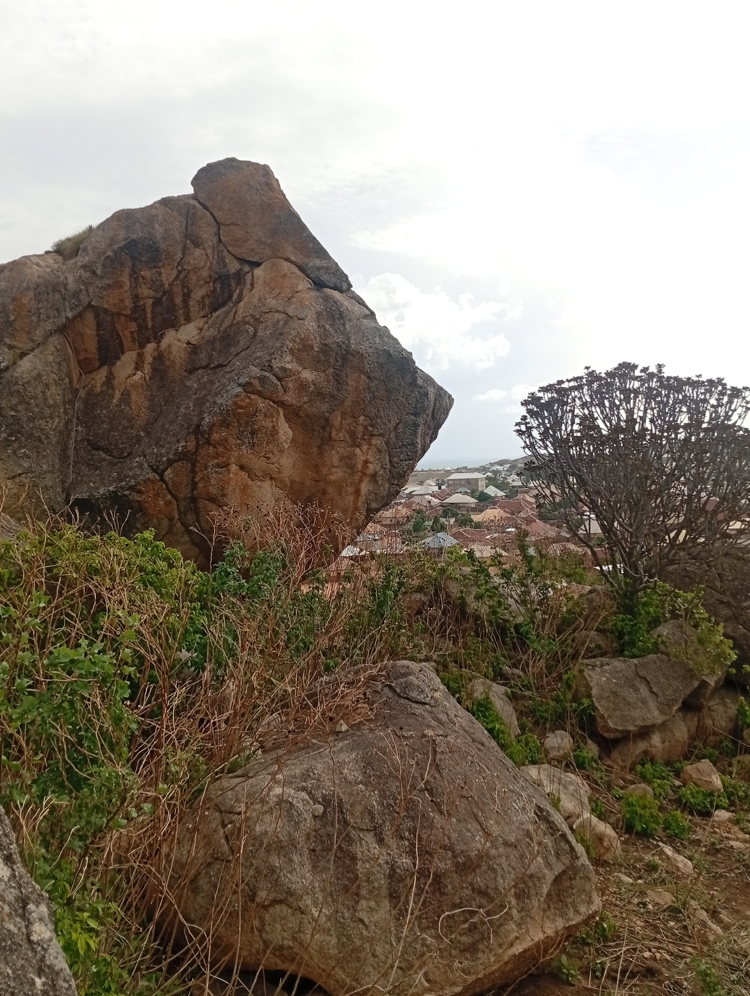 The image shows a large rock formation in Barakin Akawo, Nigeria. In the foreground, there are smaller rocks and vegetation. In the background, a section of the town is visible with buildings clustered together. The sky is overcast with scattered clouds. A tree with bare branches stands to the right of the frame.