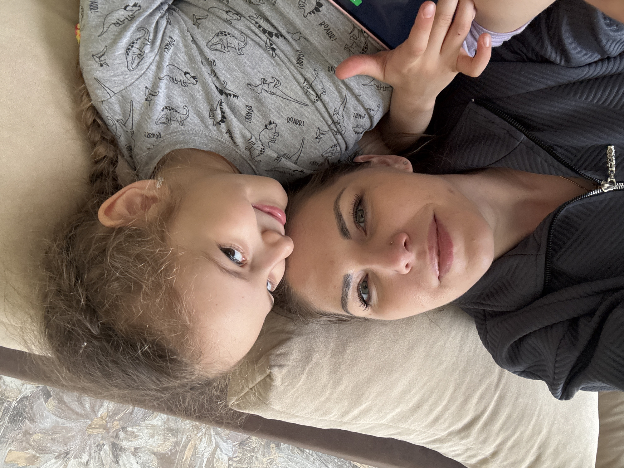A close-up, top-down image captures an adult woman and a young child lying side-by-side, heads touching. Both are looking up towards the camera, sharing a tender moment. The adult, on the right, has light eyes and dark hair, wearing a dark grey or black zippered jacket with a textured pattern. She has a subtle nose piercing. The child, on the left, has light brown, wavy hair and light eyes, displaying a wide, joyful smile. She is wearing a light grey t-shirt adorned with cartoon dinosaur illustrations and the repeated text "ROARR!". She appears to be holding a smartphone or tablet, partially visible near her hand. Both subjects exhibit calm and affectionate expressions, suggesting a loving bond. They are resting on a light-colored, upholstered surface, possibly a cushion or pillow, beneath which another patterned surface is visible. The lighting is soft and natural, indicating daytime. The scene conveys a sense of warmth and closeness.