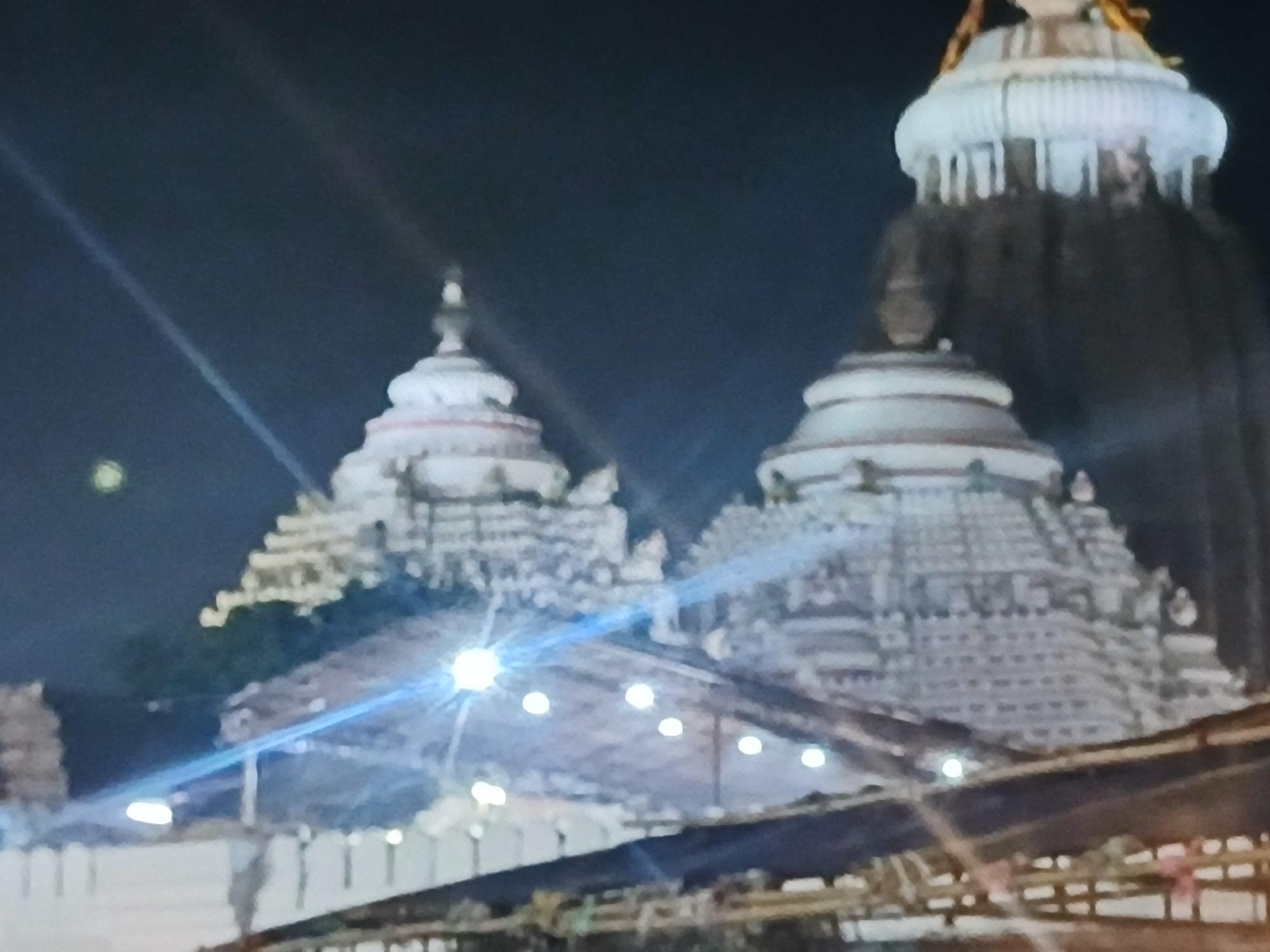 The image shows three Hindu temples at night. The tallest temple is to the right of the image, and is dark in color with a white dome. The two temples on the left are shorter, and white in color. There are bright lights shining from the bottom of the image, illuminating the temples from below. The location is likely not Bengaluru, India, as the temple architecture is not typical of that region.
