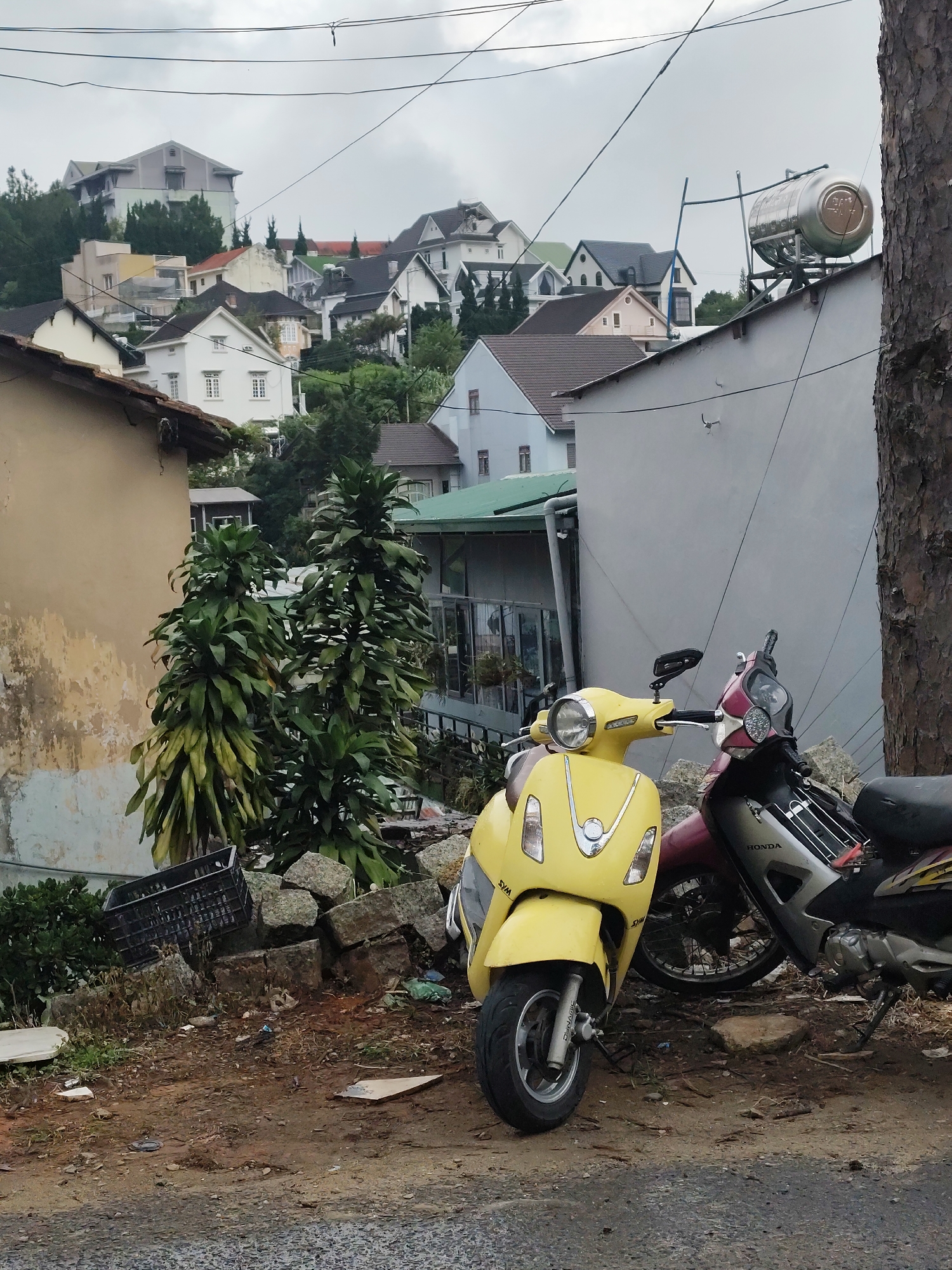 The image depicts a scene in the city of Lâm Viên - Đà Lạt, Vietnam, characterized by a hilly terrain with a dense arrangement of houses. In the foreground, two scooters are parked: a yellow scooter on the left and a red and silver scooter on the right, partially obscured. The scooters are parked on a dirt and gravel surface adjacent to a road. Behind the scooters, a light-colored building wall stands, with a tall, rough-barked tree trunk on the far right. Further back, a cluster of large green plants with broad leaves grows near a dilapidated yellow and white building. The background reveals a panoramic view of numerous houses with diverse architectural styles and roof colors, including white, gray, and red, spread across the undulating landscape. Overhead, a network of wires crisscrosses the sky, common in urban Vietnamese settings. The sky is overcast, suggesting a cloudy or potentially rainy day. Debris, including a black plastic crate and scattered stones, is visible in the mid-ground, contributing to a somewhat unkempt appearance of the immediate surroundings. A metallic water tank is mounted on the roof of a building in the middle distance.