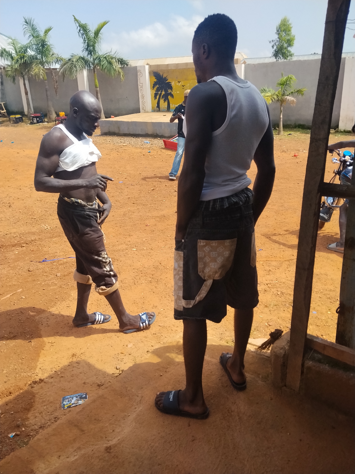 Two men are standing in a dusty, outdoor area. The man on the left is facing away from the camera, wearing dark, patterned shorts and a white bandage wrapped around his chest and abdomen. He has a shaved head and is pointing with his left hand. The man on the right is facing away from the camera, wearing denim shorts with decorative pocket flaps and a light-colored tank top. He has dark hair. In the background, a yellow and gray wall is visible with palm tree murals. There are palm trees and a building. A motorcycle is partially visible on the right side of the image. The ground is covered in orange dirt, and some debris, including a small card with images, is scattered on the ground. The scene appears to be in Durumi, Nigeria.
