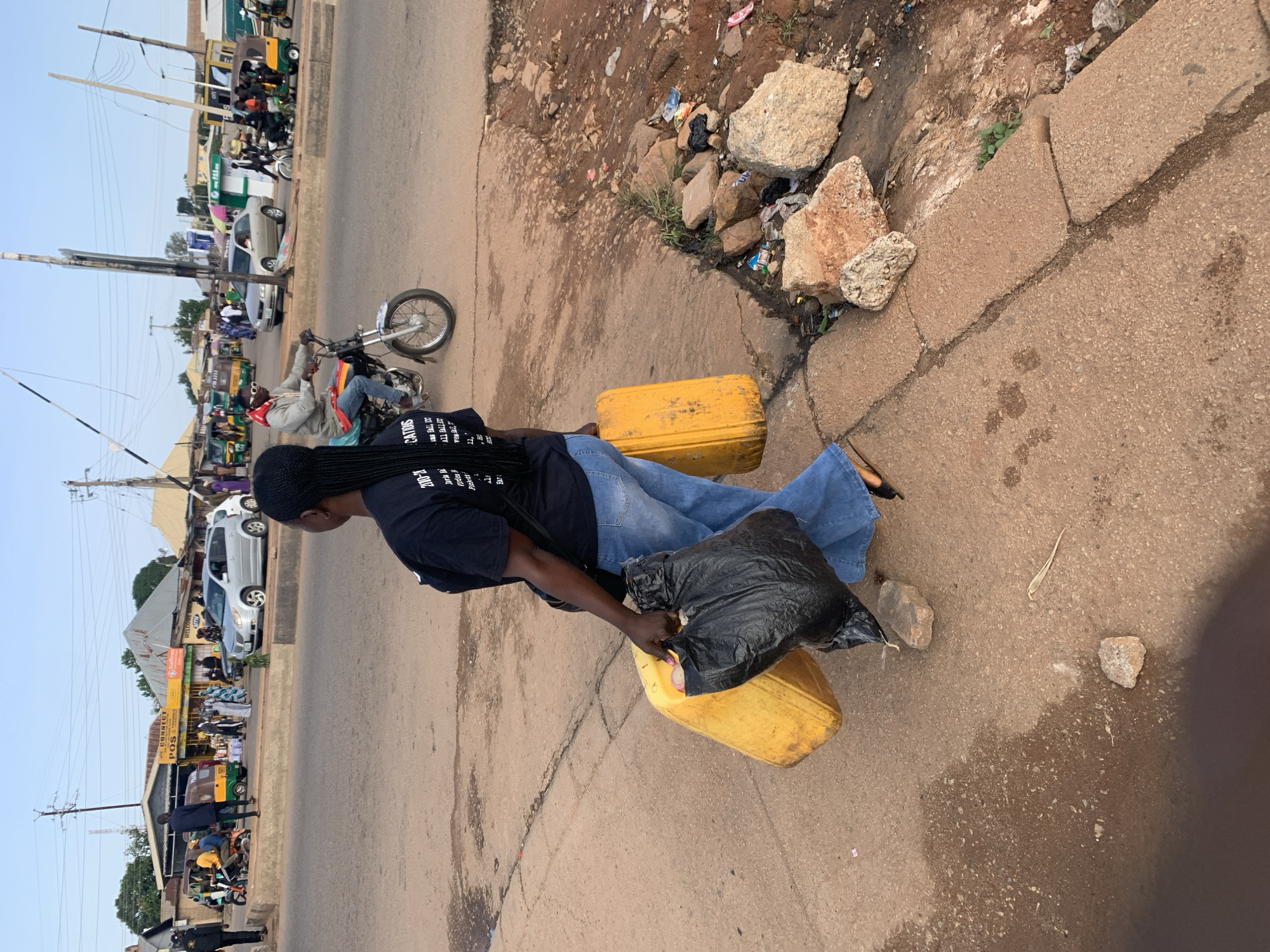 In Gura Top, Nigeria, a woman walks along a dirt road, carrying a yellow jerrycan and a black plastic bag. She is wearing a black t-shirt and blue jeans. A motorcycle passes by in the background. The scene appears to be a street in a busy marketplace, with shops and other vehicles visible. The road is made of dirt and gravel, with some debris and rocks scattered around. The sky is clear and the sun is shining.