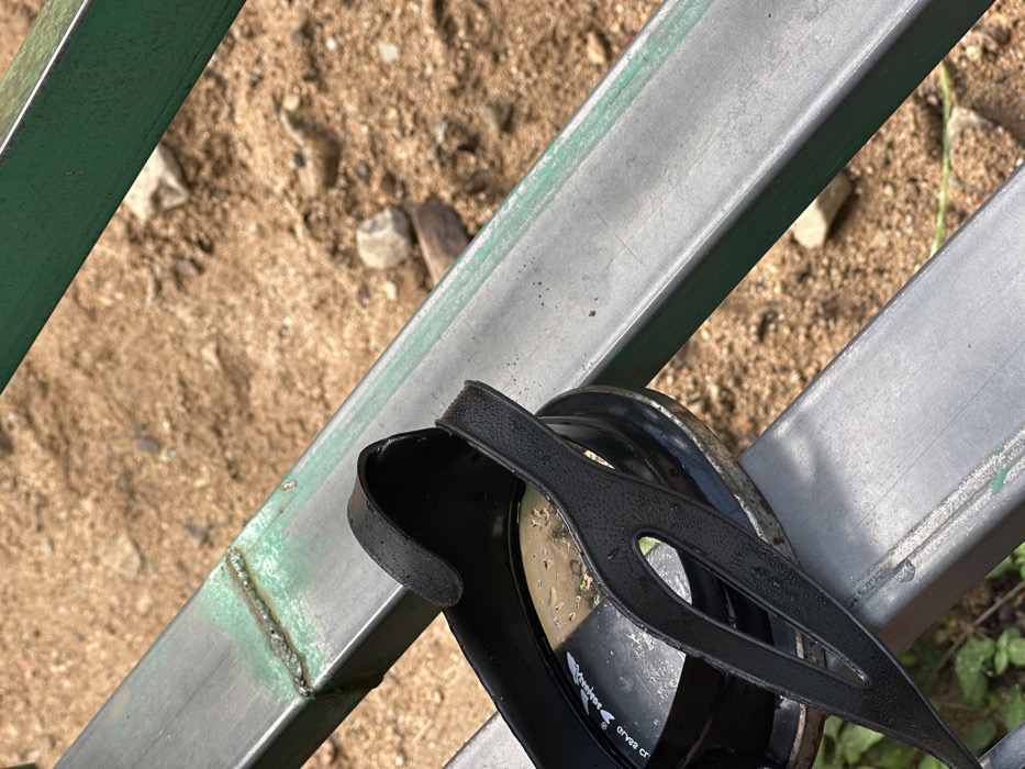 A pair of black swimming goggles rests on a weathered metal gate with green paint. The ground below is sandy with small rocks and debris. The goggles have water droplets on their straps and lenses, suggesting recent use. The gate's metal beams are worn and show signs of welding. Some green vegetation is visible at the bottom right corner.
