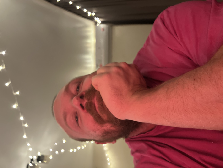 A person with fair skin, red hair, and a beard is wearing a pink t-shirt. The individual is positioned close to the camera, with their hand near their face. Behind them, string lights illuminate a room with light-colored walls. The scene appears to be indoors, possibly under a bunk bed frame. The photograph is taken at a tilted angle.