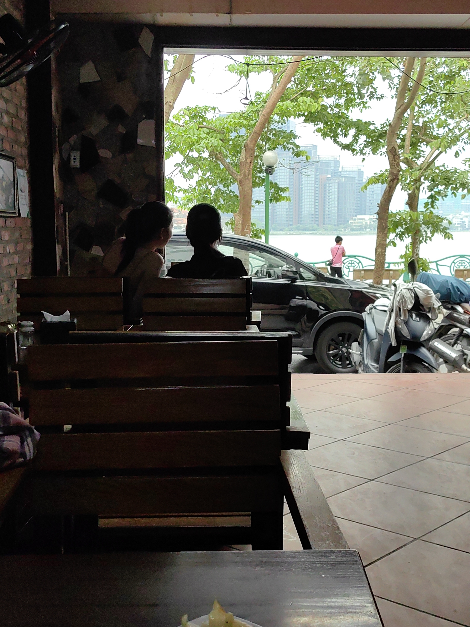 A scene from inside a building in Hà Nội, Vietnam. Two people sit on a wooden bench, looking out an open doorway or window. Outside, a black car and several motorbikes are parked near a waterfront area with a green metal railing. In the distance, tall buildings are visible across the water. A person in a pink shirt stands near the railing. The interior of the building has brick and stone walls, and the floor is tiled. A ceiling fan is visible in the upper left corner.