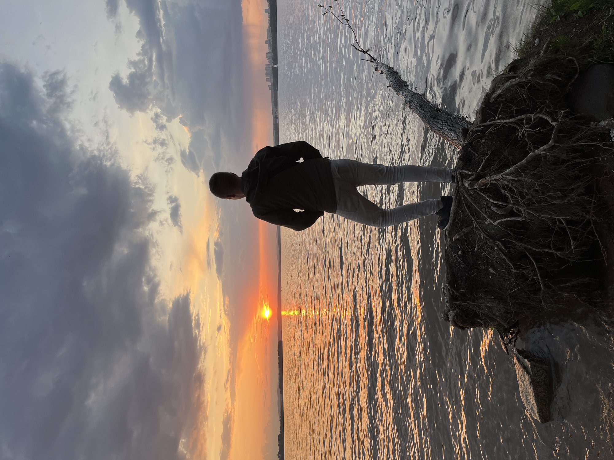 A person stands on a tree root overhanging a body of water, silhouetted against a vibrant sunset. The water reflects the orange and yellow hues of the setting sun, and a cityscape is visible in the distance. The sky is filled with clouds, some dark and others illuminated by the sunset. The location is Yekaterinburg, Russia.
