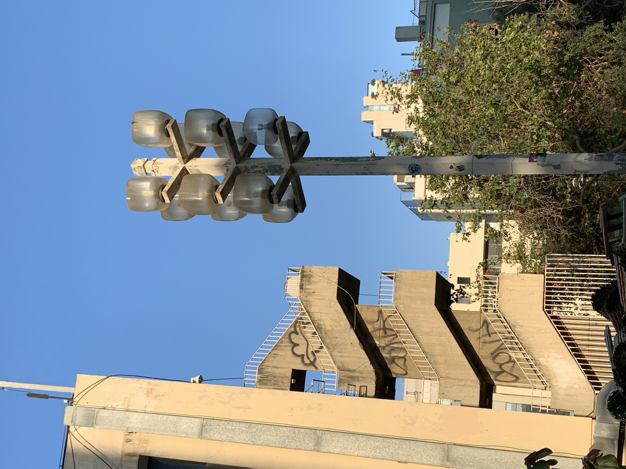 This image depicts an outdoor scene with a clear blue sky. In the foreground, a multi-tiered outdoor staircase made of concrete with white railings is visible, adorned with graffiti. To the left of the staircase is a light-colored building with gray stone accents. To the right of the staircase stands a tall lamppost with multiple glass lamp fixtures. In the background, several modern buildings and lush green trees are present. The overall setting suggests an urban environment, likely in Tel Aviv, Israel, given the architectural style and vegetation. No people are visible in the image.