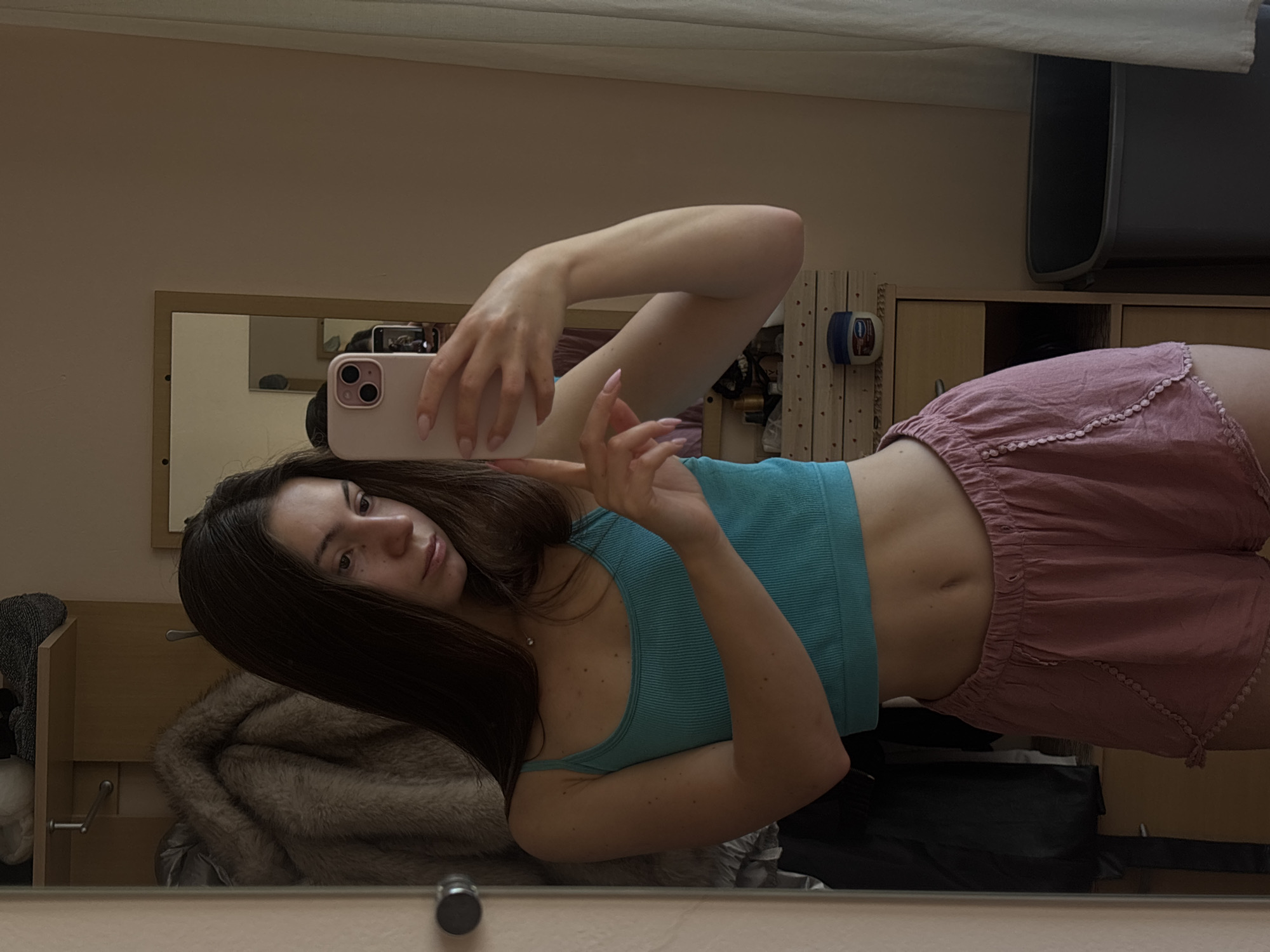 A young woman with long, dark brown hair takes a mirror selfie in what appears to be a bedroom or dressing area. She is wearing a turquoise ribbed tank top and pink shorts with decorative trim. The woman is holding a pink iPhone in her right hand, angled to capture her reflection. In the background, a mirror reflects more of the room, including a closet with a furry coat hanging inside and shelves with various items, including a small jar of Vaseline and what appears to be toiletries. To the right of the frame, a white curtain and a grey trash can are visible. The lighting suggests it is daytime. The scene is indoors, with a neutral-colored wall and wooden furniture. The location is not discernible within the city of Varna, Bulgaria, from the visual information provided.