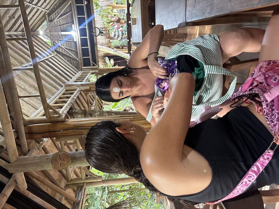 Two females are observed within an open-air, bamboo-constructed structure in Punggul, Indonesia. The female in the foreground is seen from behind, with dark hair tied in a ponytail, wearing a black tank top and gold hoop earrings. A pink and white patterned shoulder bag rests on her left side. She extends her right arm towards a large white and green striped tote bag. The second female, partially obscured and facing away from the viewer, has wet, dark hair with a white oval marking on her forehead and wears a dark-colored top. She is actively reaching into the striped tote bag, which contains various colored fabrics including purple, green, and black. The surrounding environment consists of bamboo structural elements, including columns, beams, and a slatted roof. A carved wooden mask is affixed to a bamboo column in the upper left. In the background, lush green tropical vegetation is visible, alongside additional wooden tables and decorative elements, including dark, horn-like hanging objects and patterned textiles. The scene is illuminated by ambient daylight.
