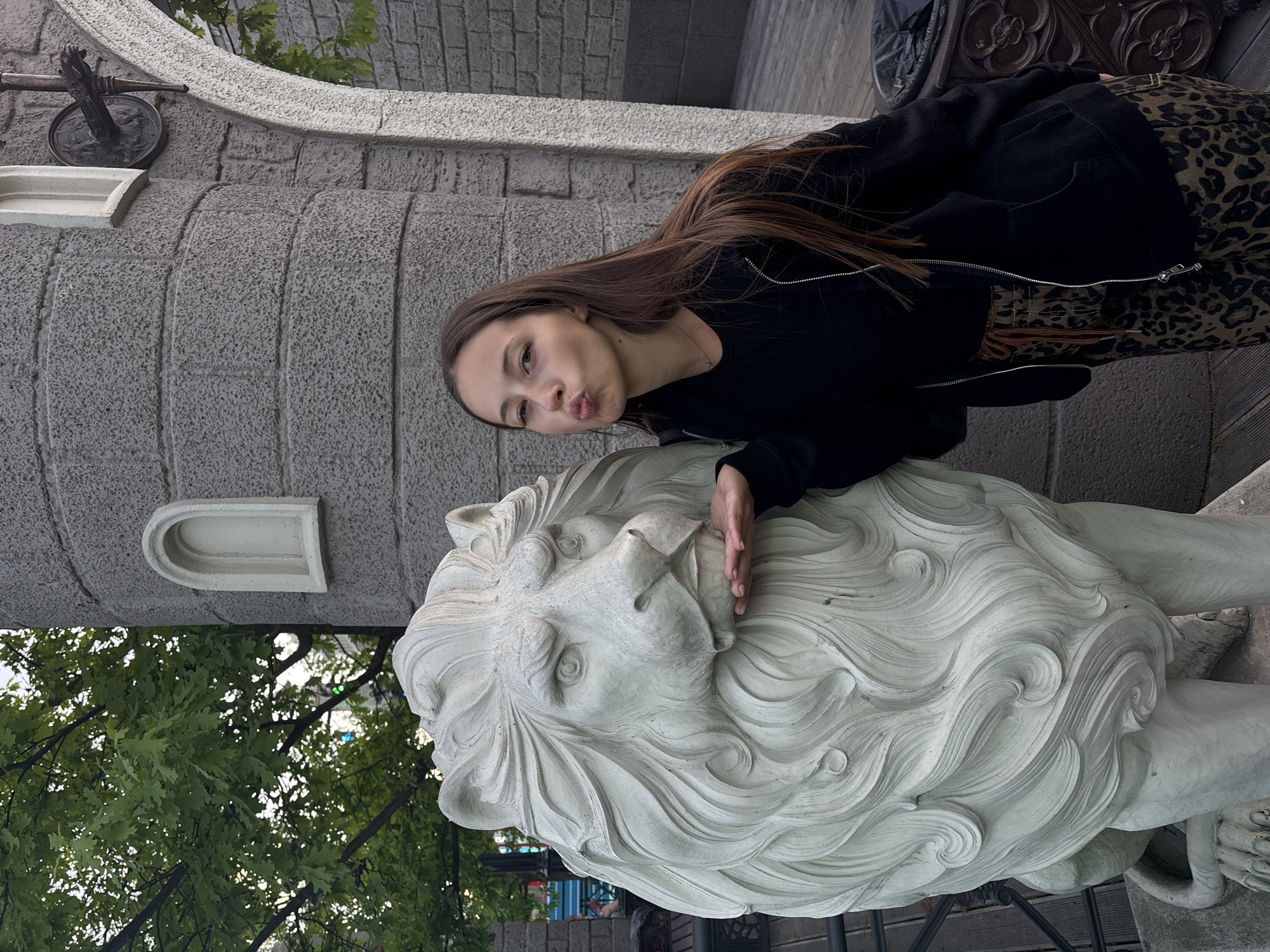 A young woman with long brown hair poses with a statue of a lion. She is wearing a black jacket and leopard print pants. She is leaning against the lion statue and making a kissing face. The lion statue is made of white stone and has a detailed mane. In the background is a grey stone building, possibly a castle. The location is Moscow, Russia.