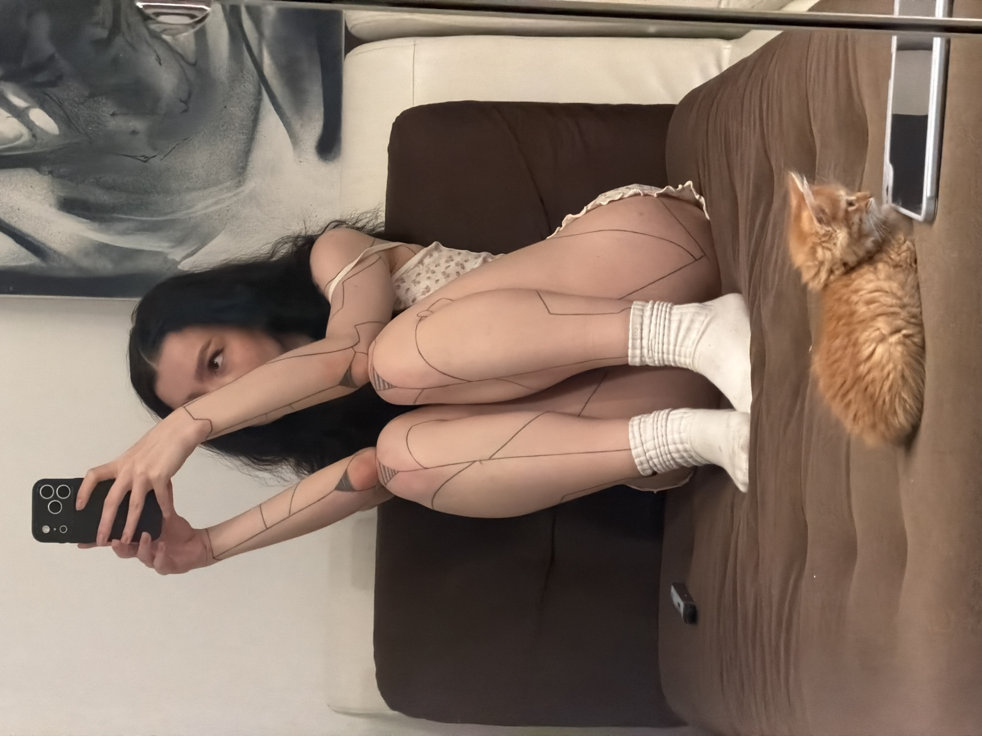 A woman is lying on a brown sofa, taking a selfie with a black iPhone. An orange cat is standing on the sofa next to her. The background includes a white cushion, a black and white painting and a chrome bar. The scene appears to be indoors. It is not possible to verify location in Khimki, Russia from visual data.