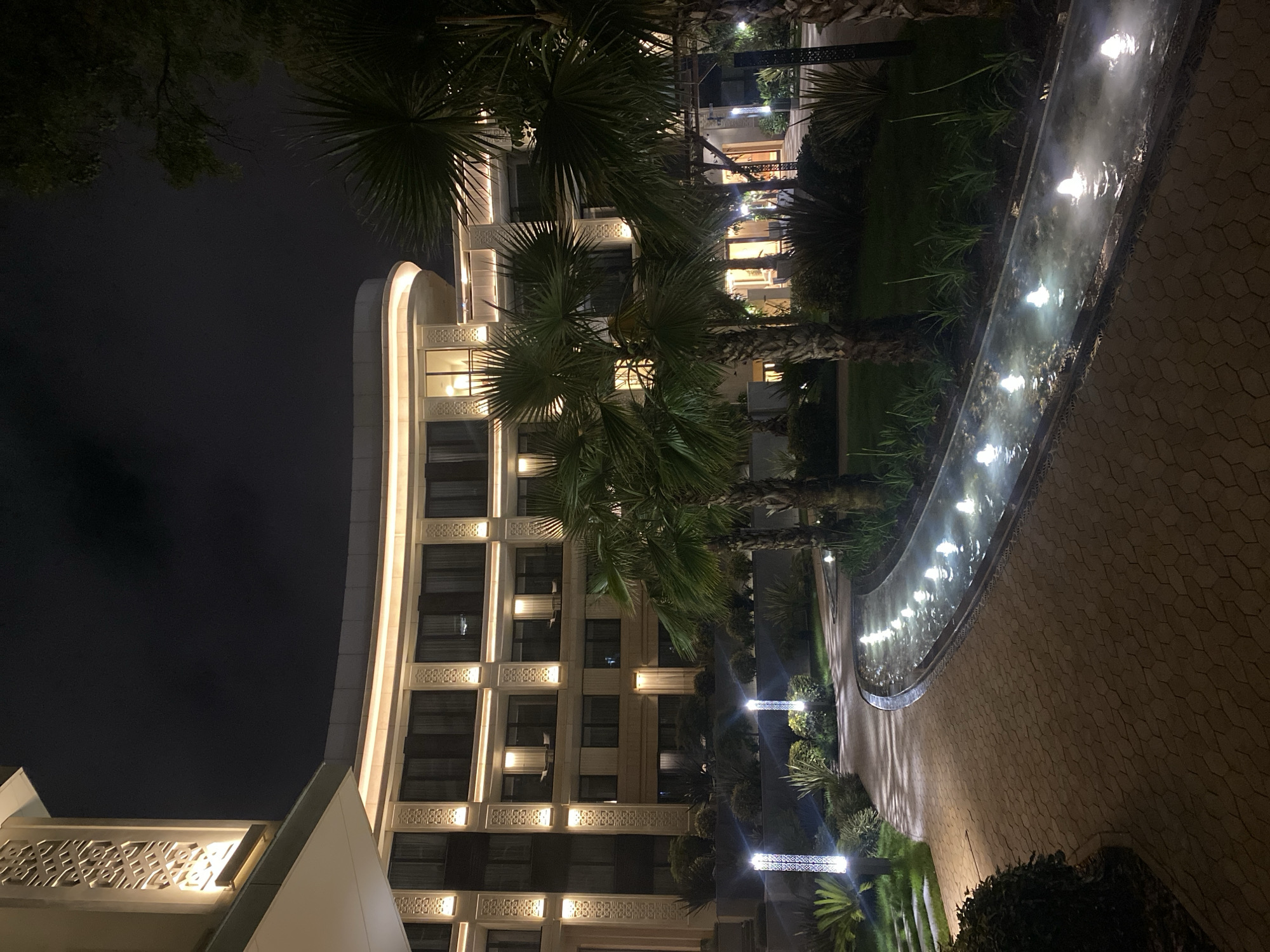 This image depicts the nighttime exterior of a modern hotel in Sochi, Russia. A curved, multi-story building with illuminated decorative panels and numerous windows is visible. The building is lit by warm, upward-facing lights along its edges and vertical accent lights on its facade. In the foreground, a pathway paved with hexagonal stones curves to the right, leading past a narrow, linear water feature with multiple spouts creating illuminated fountains. Palm trees and lush green vegetation surround the pathway and water feature. The sky is dark and cloudy, indicating nighttime.