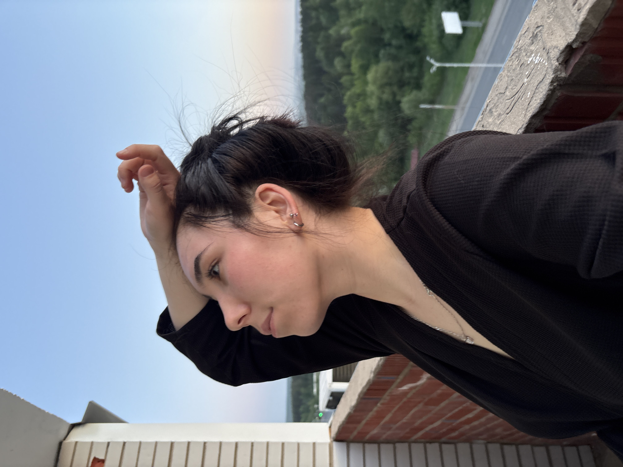A young woman with dark hair pulled into a bun is resting her forehead on her hand while leaning over a brick and concrete ledge. She is wearing a black top and a silver necklace. Her ear is pierced with multiple silver earrings. In the background, a blurred landscape reveals green trees and a light-colored sky. To the right, there is a gray road with street lights. The ledge appears to be part of a building structure with a tile facade. Without location information, it is not possible to identify the city as Izhevsk, Russia.