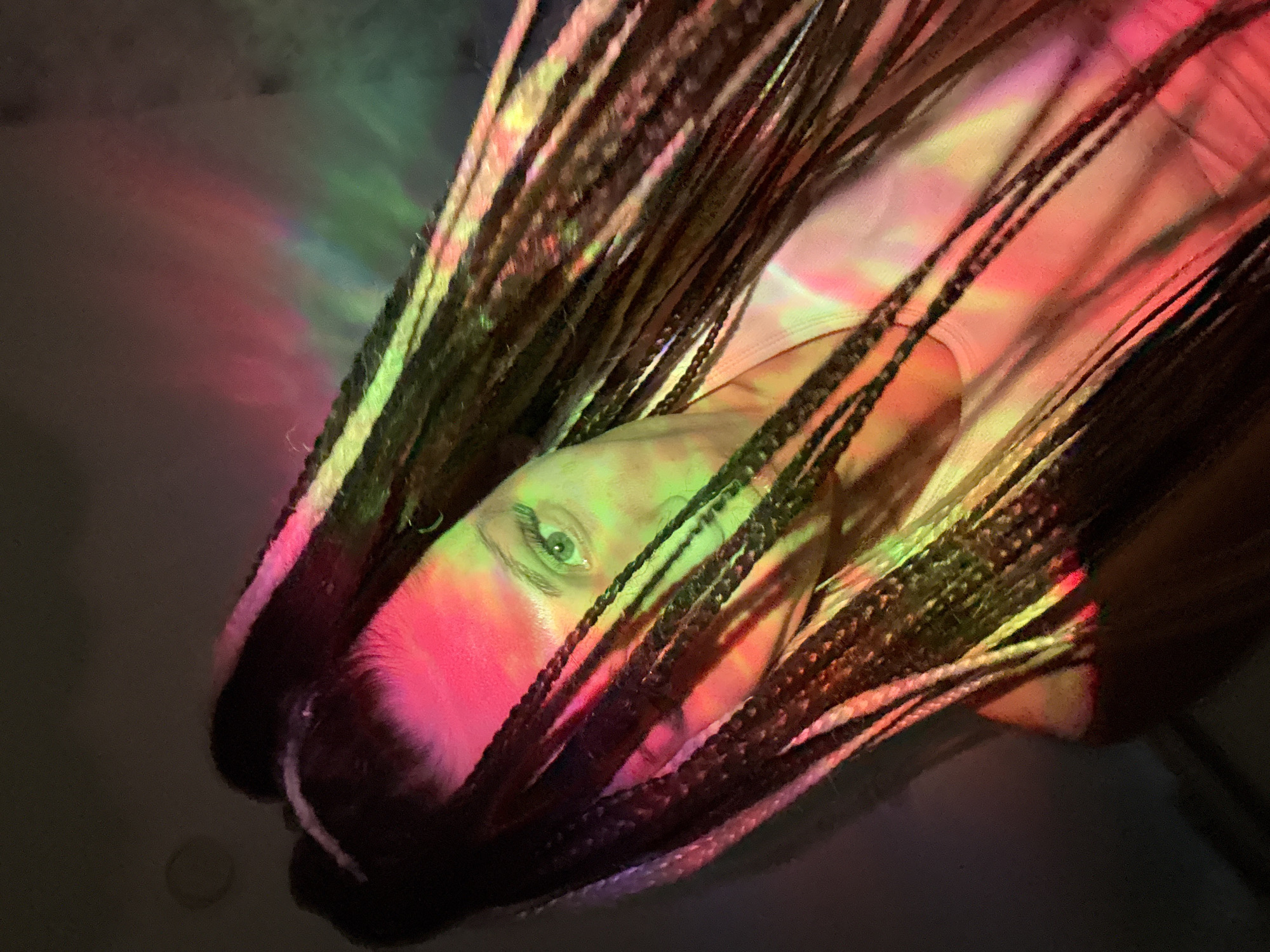The image displays a close-up, angled portrait of a person with long, dark braided hair, possibly dreadlocks. The individual's face is partially obscured by the braids, with one eye (the subject's right eye, located on the left side of the frame) clearly visible and appearing green. The scene is characterized by dramatic and colorful artificial lighting, casting prominent hues of pink/red and green, with subtle blues and purples, across the subject's face and hair. These light beams create dynamic streaks and patterns. The background is dark and indistinct, suggesting an indoor setting with low ambient light. The person appears to be looking towards the camera. The mood is artistic and somewhat ethereal due to the vibrant lighting. No specific activity is taking place beyond posing for the photograph. There is no visible text. The time of day is implied to be evening or night due to the reliance on artificial lighting. The image provides no specific visual cues to its stated location of Bor, Russia.