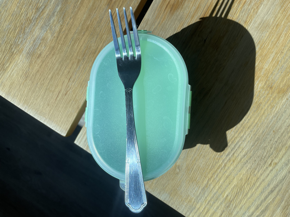 The scene depicts a light green, oval-shaped plastic lunch box with a translucent lid, positioned centrally on a light-colored wooden surface. Resting horizontally on the lunch box lid is a silver metal fork with four tines and a decorative handle featuring parallel ridges. Strong direct sunlight illuminates the objects, casting prominent dark shadows from both the lunch box and the fork onto the wooden surface, extending towards the lower right. A large, dark, undefined shadow occupies the upper left portion of the image. The wooden surface displays a visible grain pattern. The location is Montigny-le-Bretonneux, France. No individuals are present.