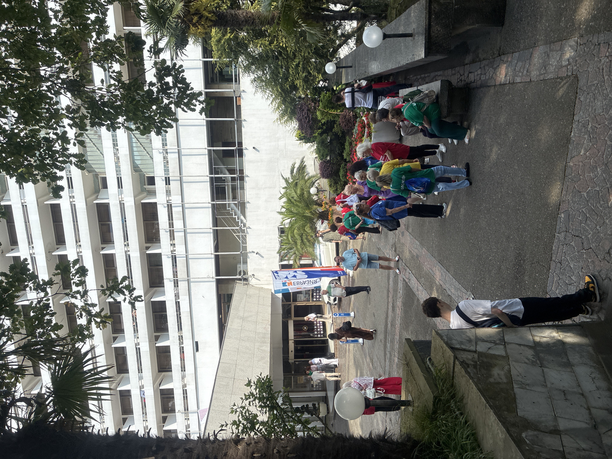 The image depicts a group of people gathered outside a large, white, multi-story building with numerous balconies, likely a hotel or resort, with the surrounding vegetation suggesting a temperate climate. Several individuals are standing in conversation or observing their surroundings, while others are seated on a stone bench. The building appears to be a modern architectural design. A white car is parked near the entrance of the building, and a large banner with text is displayed. The overall scene suggests an outdoor event or gathering. The presence of palm trees and lush greenery in the foreground and background implies a location with a pleasant climate. The architecture of the building, with its repetitive balcony structures, is characteristic of many resort hotels. The lighting suggests it is daytime, possibly late morning or early afternoon, with sunlight casting shadows. The people are dressed in casual attire, and some appear to be wearing t-shirts or uniforms, hinting at a potential organized activity or conference. The presence of outdoor lamps on posts suggests that the area is illuminated at night. The ground is paved with a mix of asphalt and decorative stone. In the foreground, a young man in a white t-shirt and dark pants with a backpack is leaning against a stone wall. Further back, a cluster of people are standing near the entrance, some in conversation, others looking towards the building. The atmosphere appears relaxed and social.