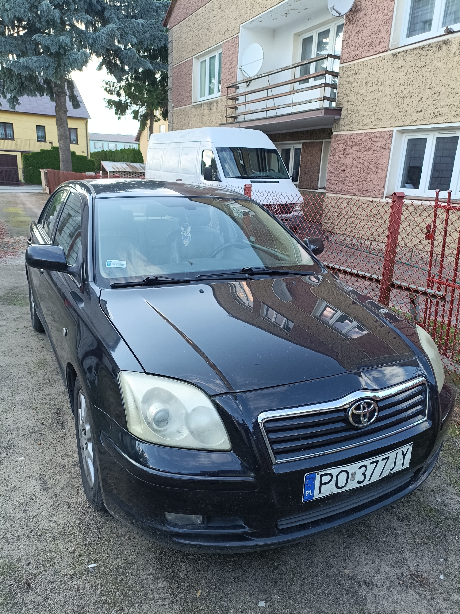 A black Toyota sedan with license plate PO 377JY is parked on a gravel surface in front of a multi-story building in Ciechanowiec, Poland. A white panel van is parked behind the sedan. The building has a textured facade with light brown and pink sections and several windows. A red chain-link fence with metal posts runs in front of the building. Tall evergreen trees are visible in the background, along with other residential buildings. The sky is overcast.