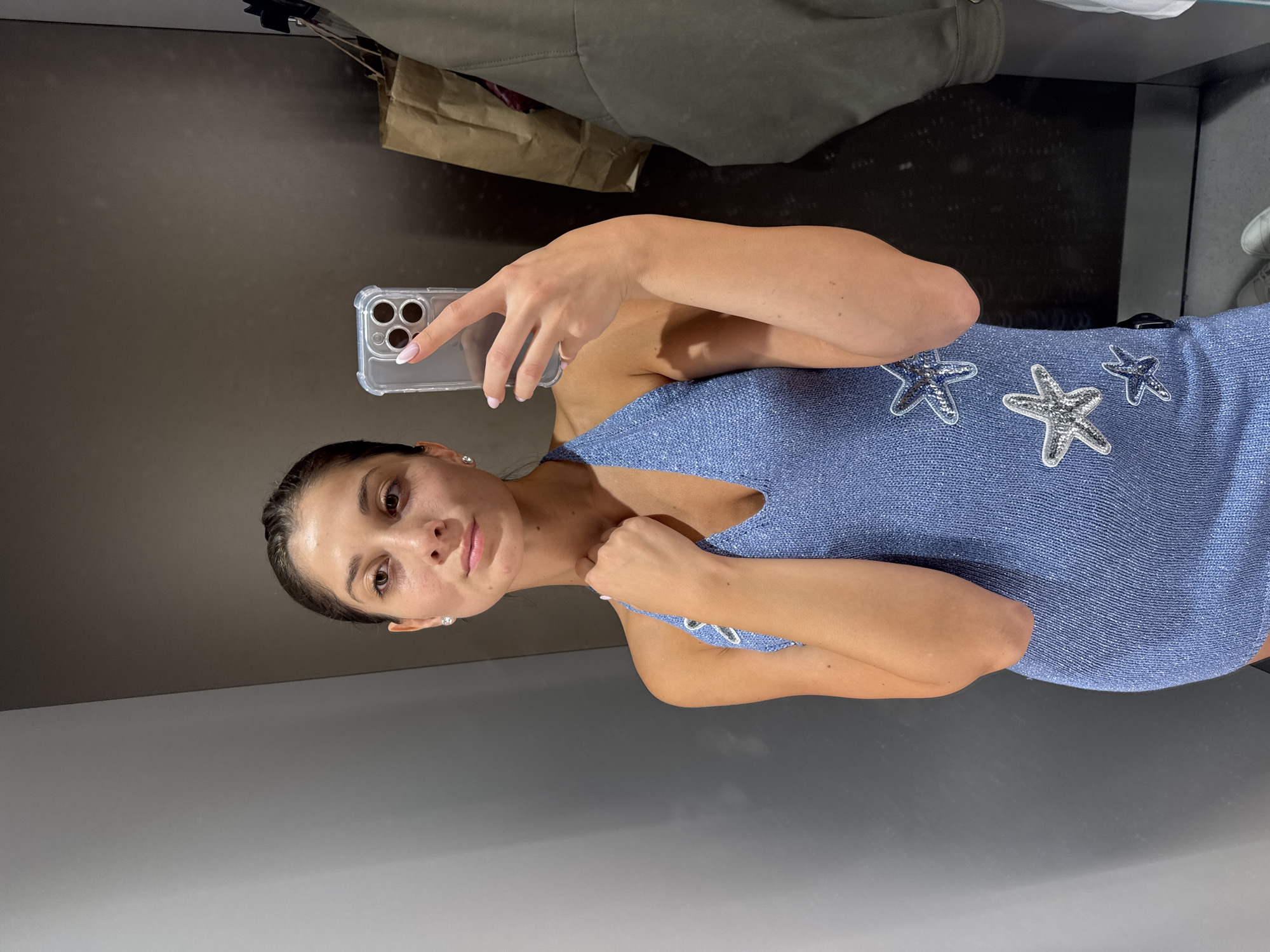 A young woman is depicted taking a mirror selfie in what appears to be a fitting room or a retail store in Burgas, Bulgaria. She holds a smartphone, equipped with a clear case and visible camera lenses, in her right hand, with her fingers positioned to operate the device. Her fingernails have a neat, light-colored, possibly French manicure style. Her dark hair is neatly pulled back, and she wears small, sparkling stud earrings. She looks towards the phone's reflection with a calm, neutral expression.
She is wearing a light blue, textured, sleeveless top or short dress that features a V-neckline. The garment is adorned with several prominent, silver, glittery starfish embellishments on the chest and shoulder area. The background of the mirror reflection shows plain, dark grey walls. Above her shoulder, reflections of hanging clothing, including a dark green or grey jacket-like item, and a brown paper bag are visible. A white sneaker can also be partially seen in the reflection. The scene is brightly lit, typical of an indoor retail environment. No visible text is present in the image.