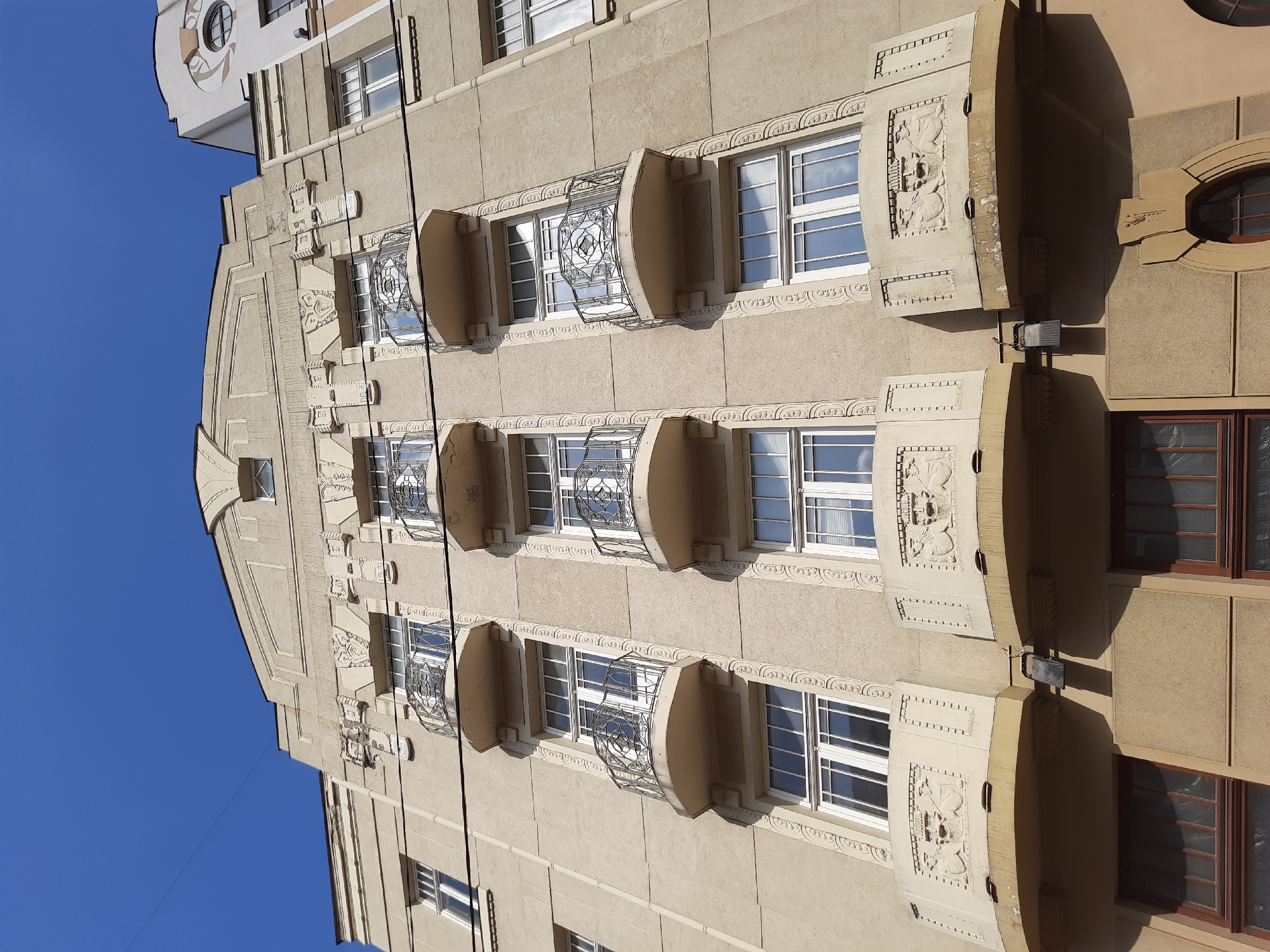 This is a low-angle, full shot of the facade of a tan-colored building in Lviv, Ukraine. The building has a distinct Art Nouveau architectural style, characterized by its curved lines, decorative elements, and ornamental balconies. The facade features three vertical rows of windows, with each row punctuated by three balconies on different levels. The balconies are circular and supported by ornate brackets. The railings of the balconies are made of decorative wrought iron with intricate patterns. The lower sections of the balconies are adorned with bas-relief sculptures depicting human faces and other decorative motifs. Above the windows, the building's facade is decorated with geometric patterns and stylized floral elements. The sky above is a clear, bright blue, suggesting a sunny day. There are power lines visible against the sky, crossing the upper portion of the building. To the right, a portion of an adjacent building with a different architectural style is visible, featuring a circular window with decorative elements. The building's lower floors have darker windows with wooden frames. The scene is devoid of any people or visible activity.